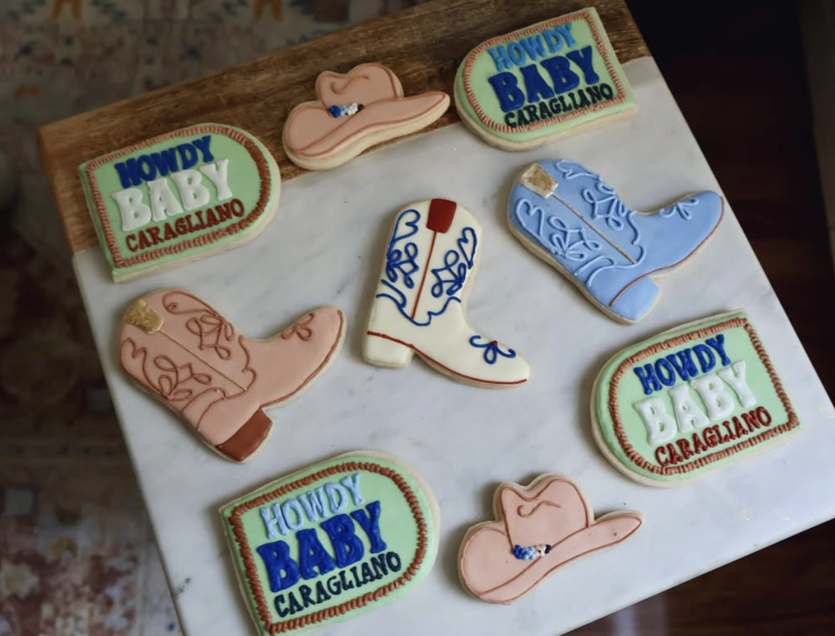 Decorative cookies with cowboy hat shapes, boot shapes, and square signs reading 'Howdy Baby Caragliano' on a white marble surface.