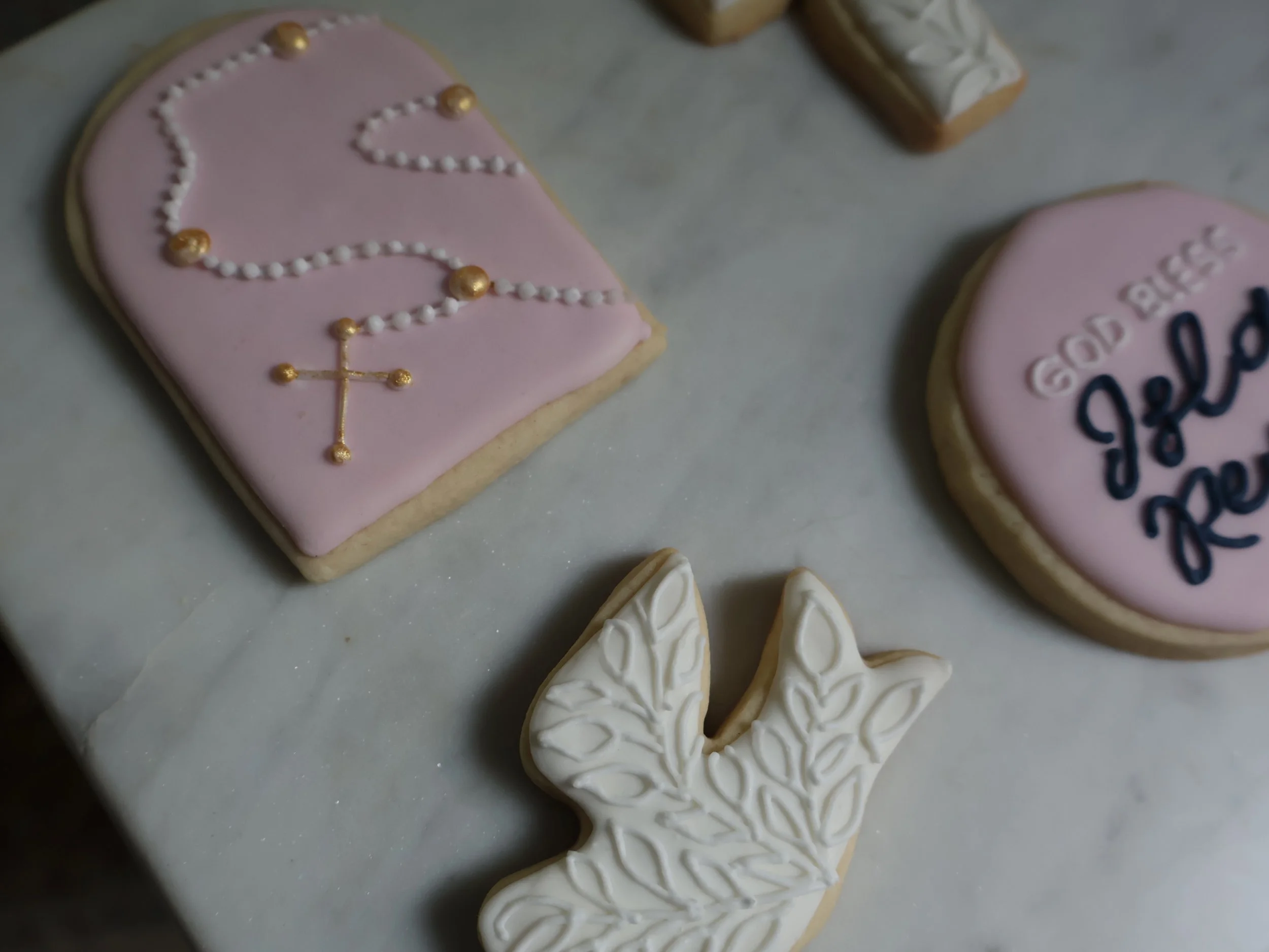 Decorated cookies on a white surface, including a pink cookie with a rosary design made of gold and white icing, a pink circular cookie with scripture text, and a white dove-shaped cookie with leaf patterns.