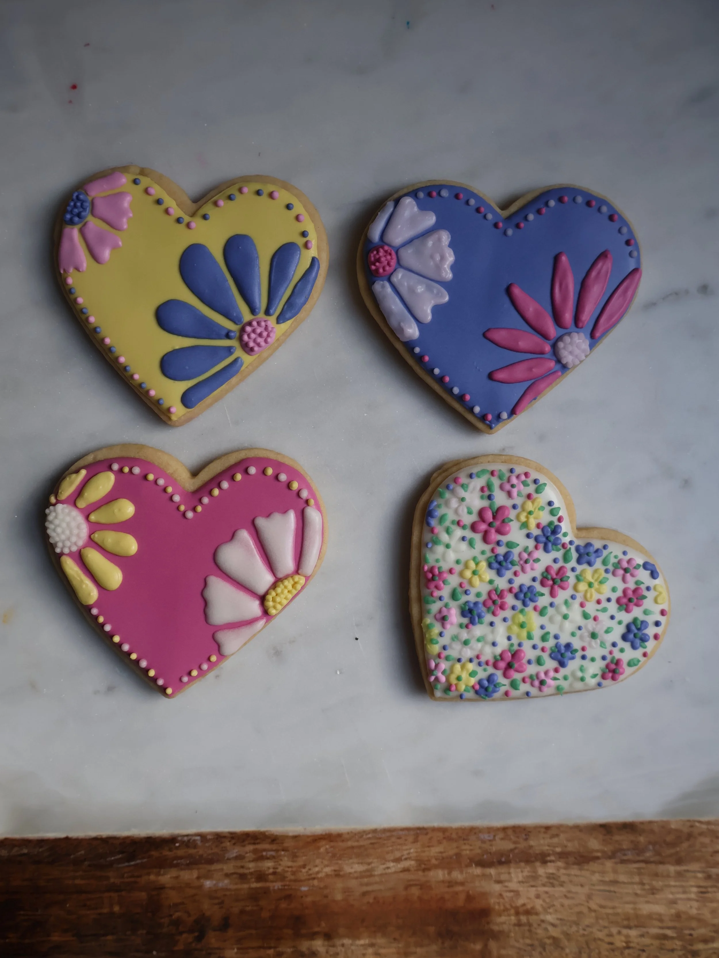 Four decorated heart-shaped cookies with colorful floral icing patterns on a light surface.