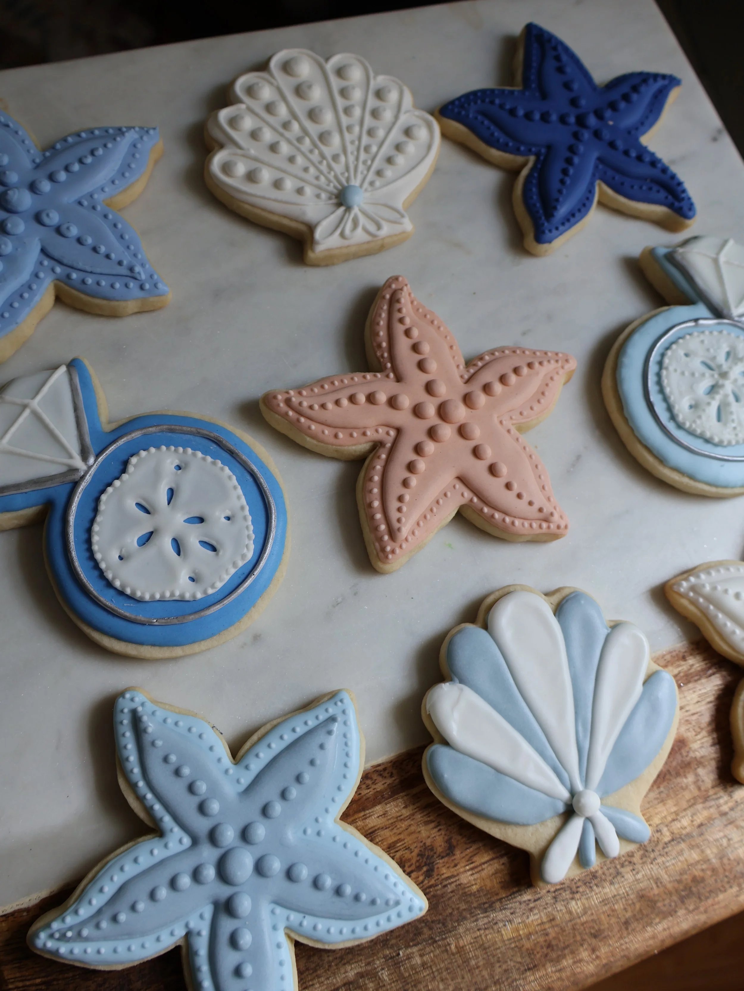 Decorated starfish, seashell, and nautical-themed sugar cookies in blue, pink, and white icing on a marble surface.