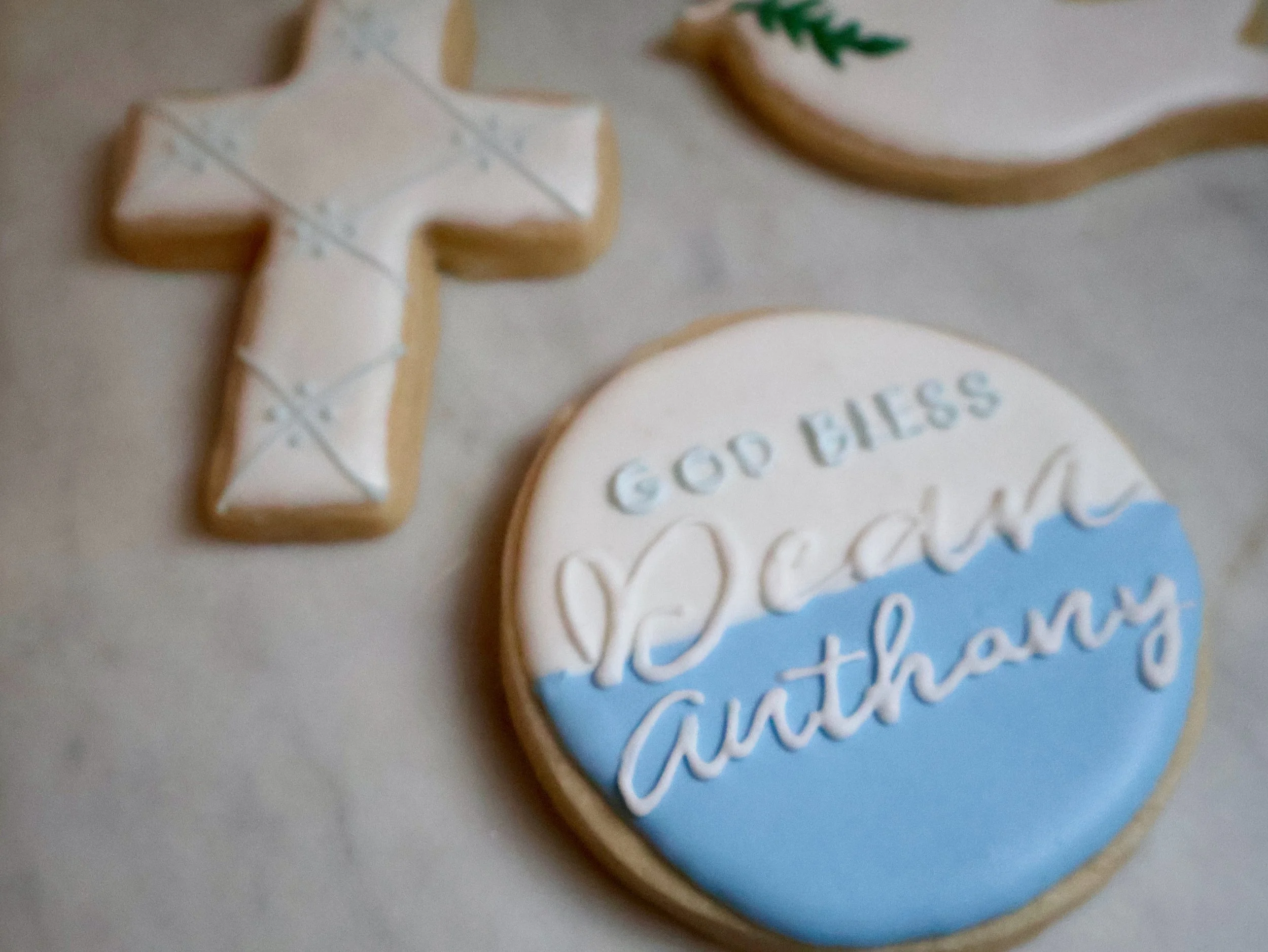 Decorative cookies, one shaped like a cross with white icing and blue decorative lines, and another round cookie with white, blue, and white icing that says 'Good vibes beachy anytime.'