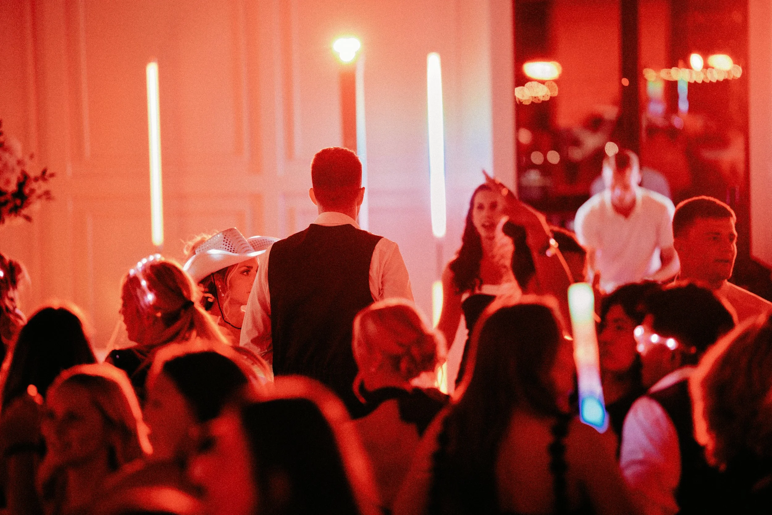 People at a lively party or social event with red lighting, some dressed in costumes or fancy attire, standing and talking in a crowded room.
