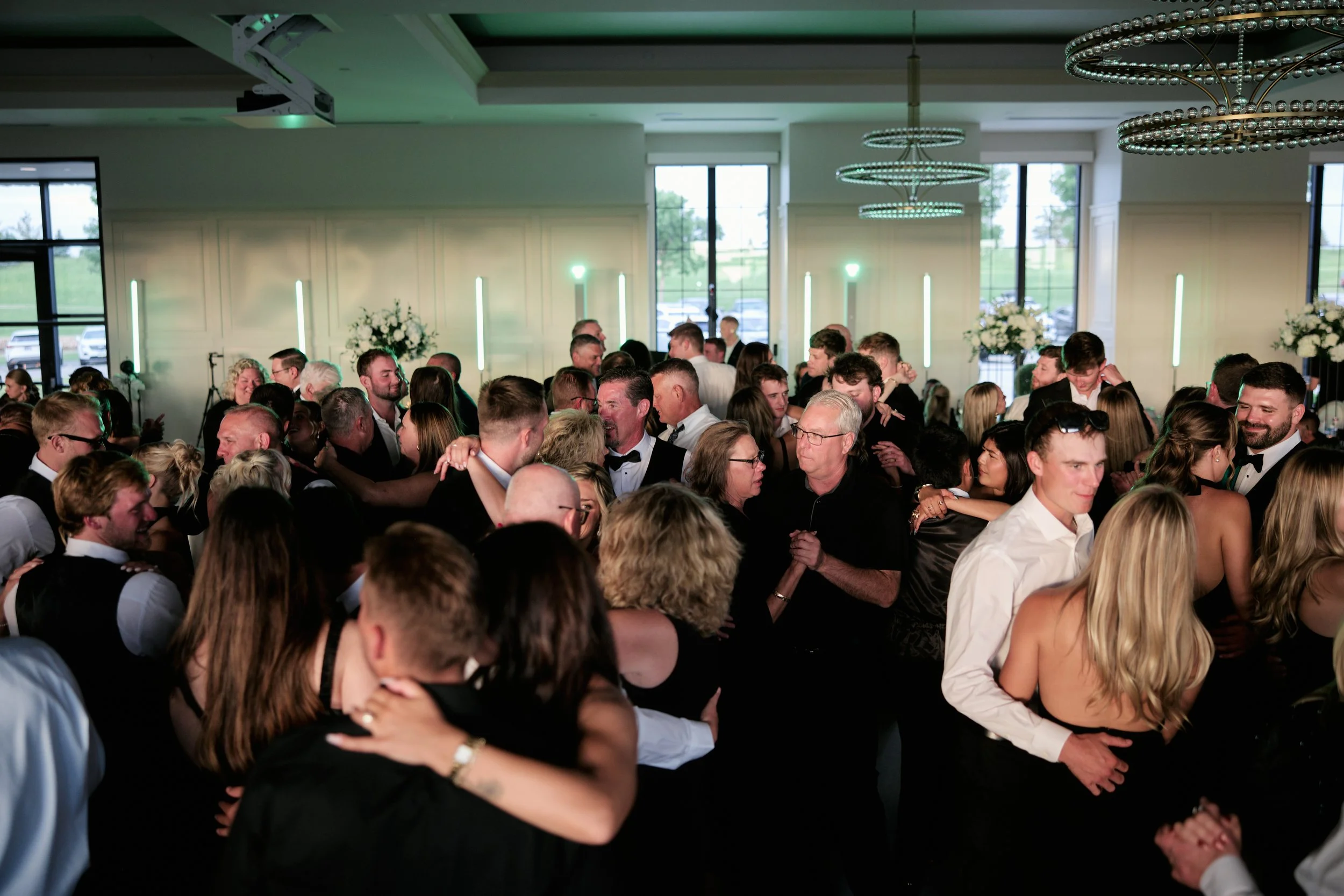 A crowded indoor dance floor filled with people dancing and smiling at a celebration or party, with large windows, floral arrangements, and modern light fixtures.
