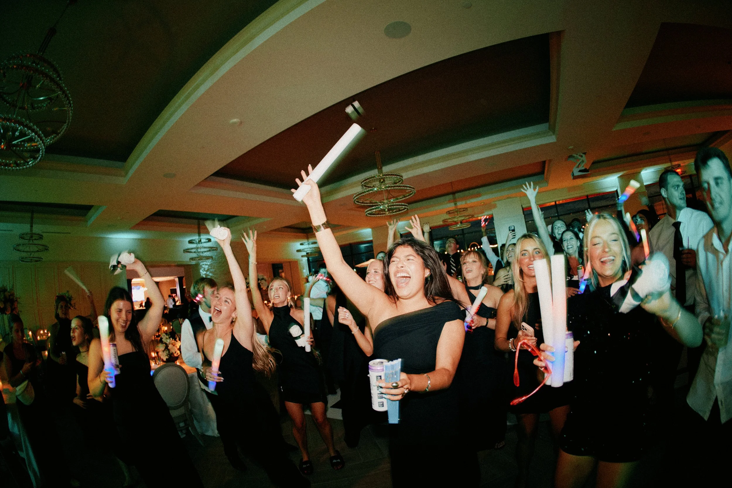 A group of people celebrating at a party or event, dancing and holding party poppers and glow sticks in a banquet hall with dim lighting and decorative ceiling fixtures.