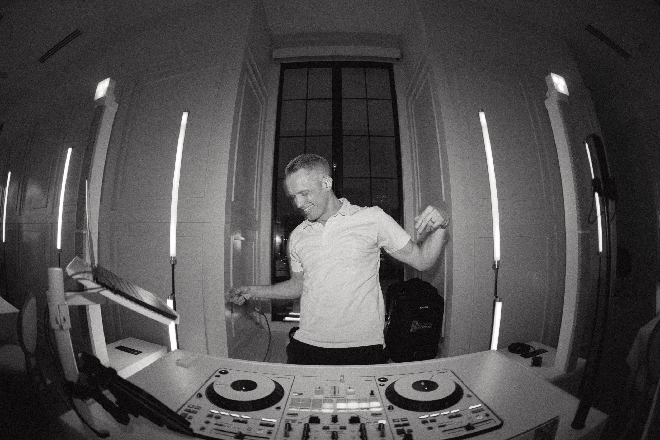 A man smiling and dancing behind a DJ booth with turntables and equipment, illuminated by vertical light bars in a room with high ceilings and large windows.