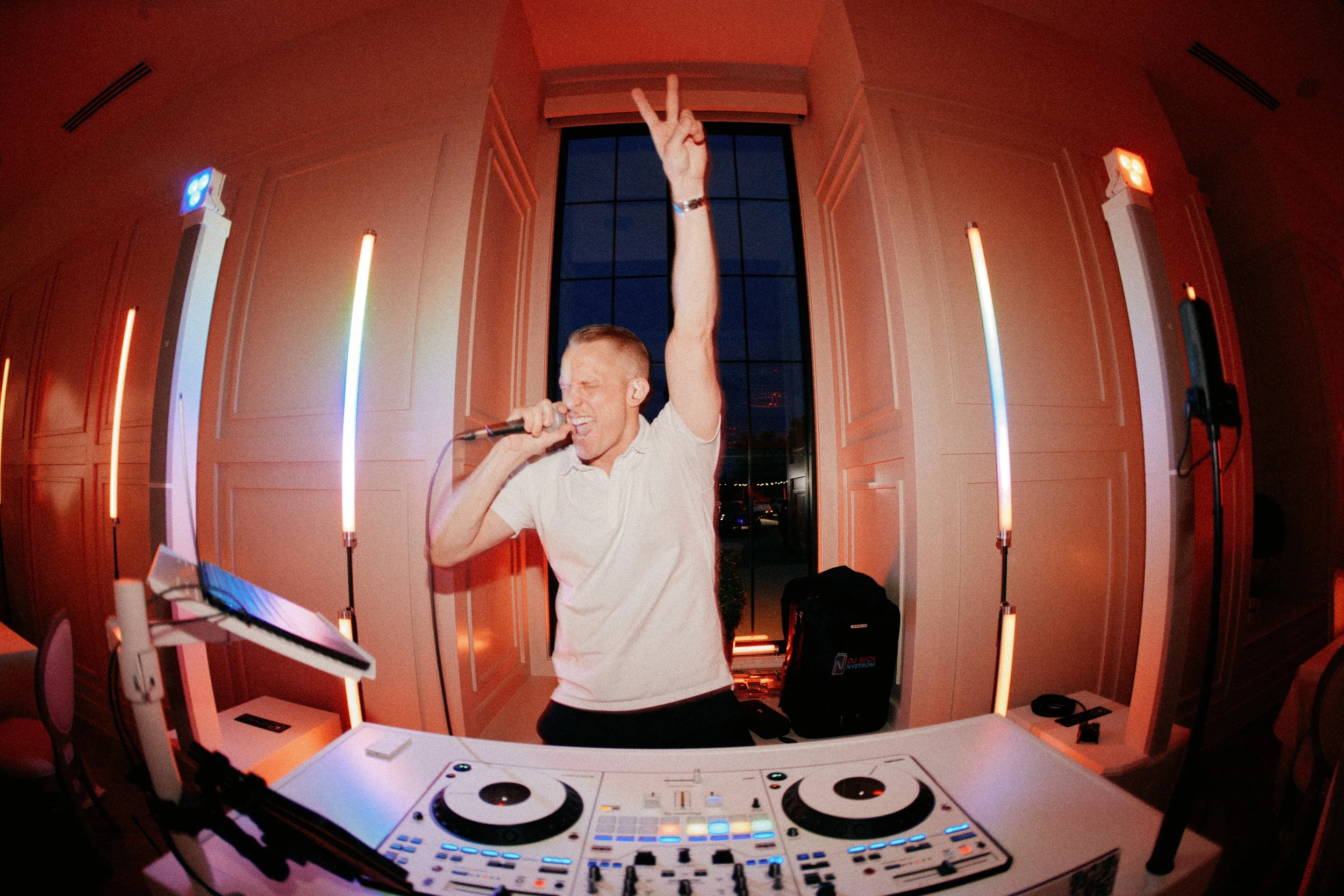 A young man singing and raising his hand with a peace sign at a DJ setup in a room with pink and blue lighting.