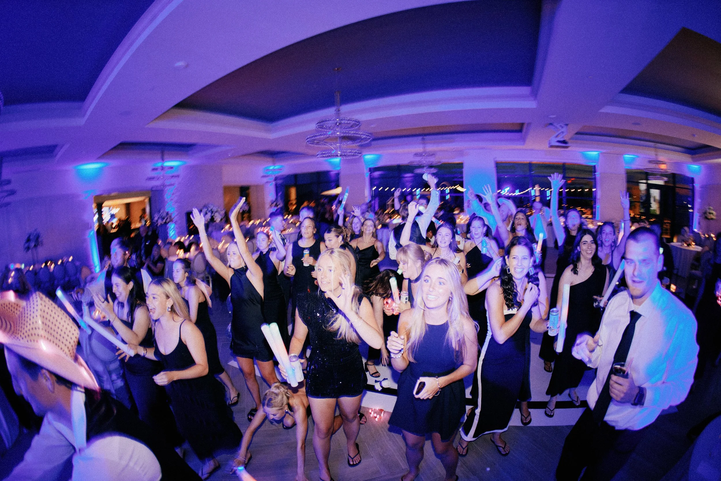 People dancing and enjoying themselves at a lively indoor party with blue and purple lighting, chandeliers, and large windows.