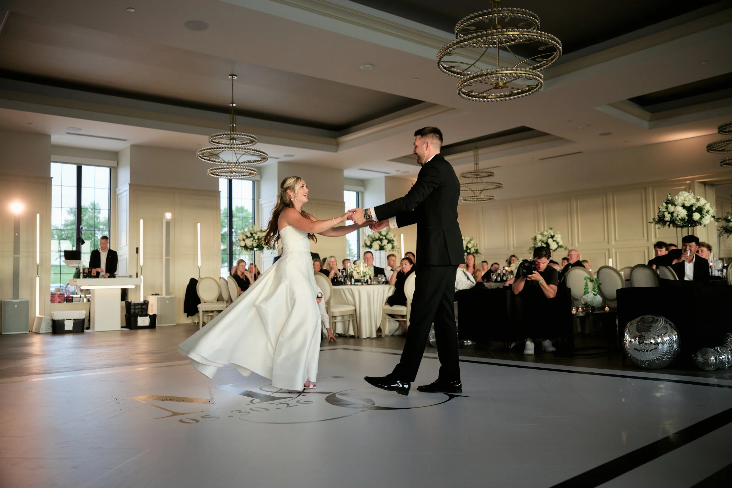 A bride and groom dancing at their wedding reception in a decorated banquet hall with guests seated at tables, floral arrangements, and modern chandeliers.