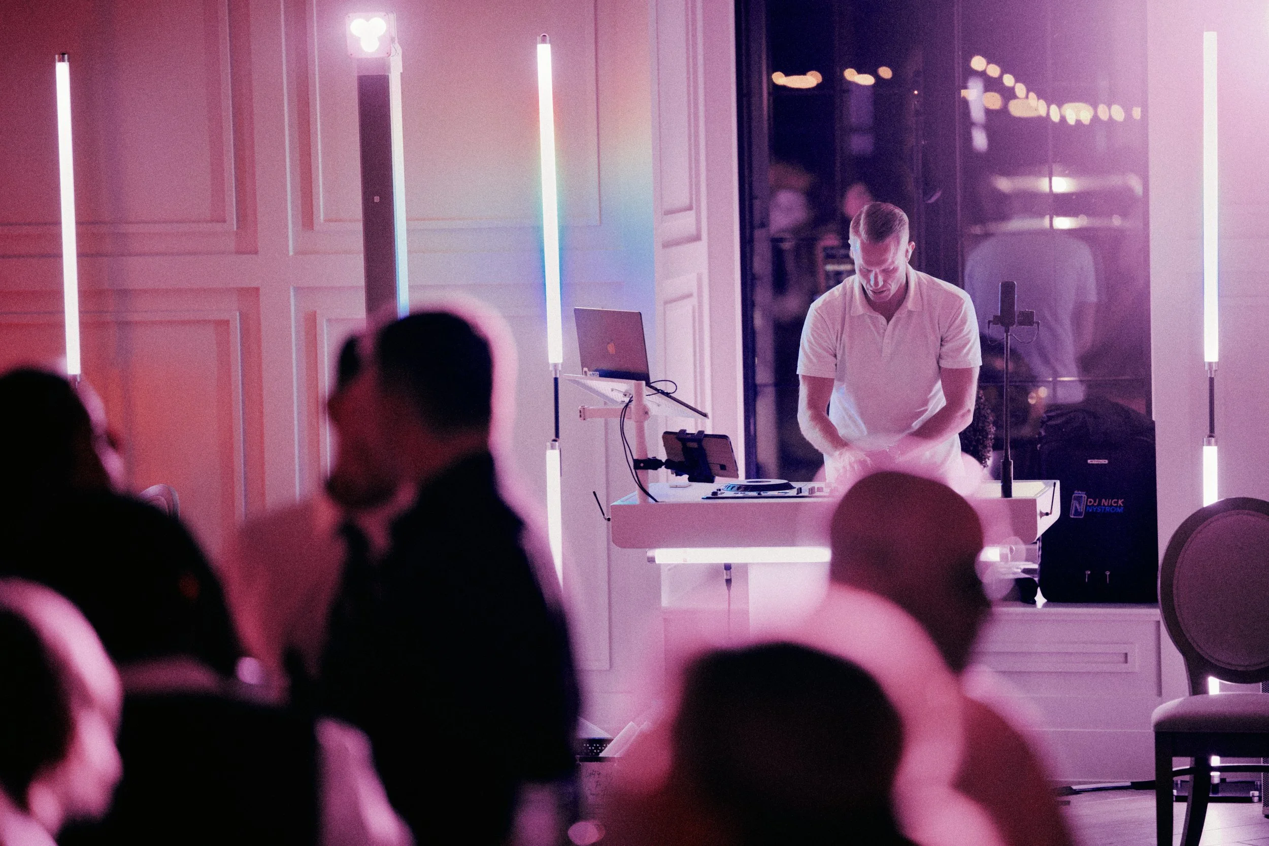 A DJ performing at an event with people seated and watching. Colorful vertical light bars illuminate the scene.