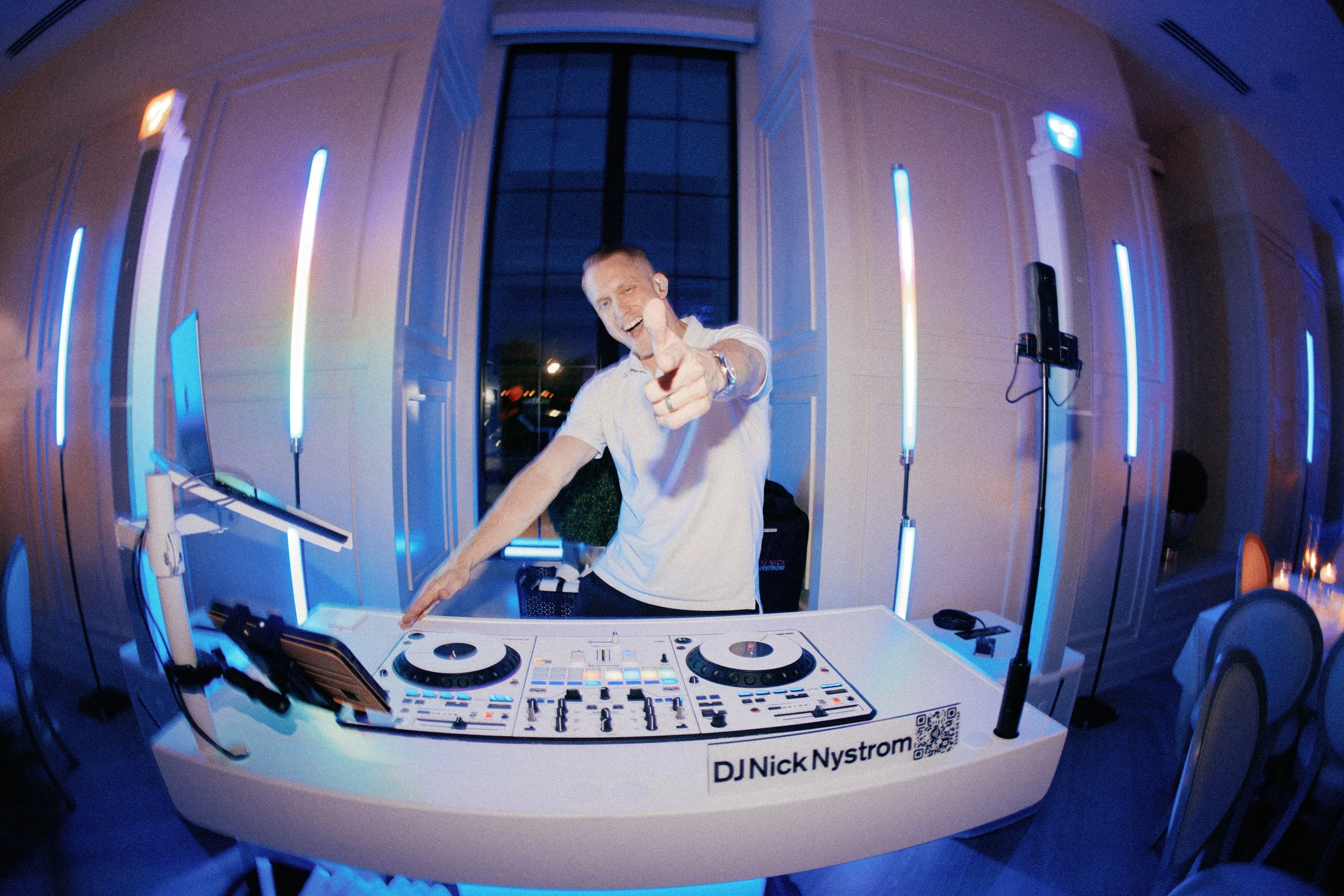 A man standing behind a DJ controller, smiling and pointing at the camera, in a room with blue LED lighting, with DJ equipment labeled 'DJ Nick Nystrom'.