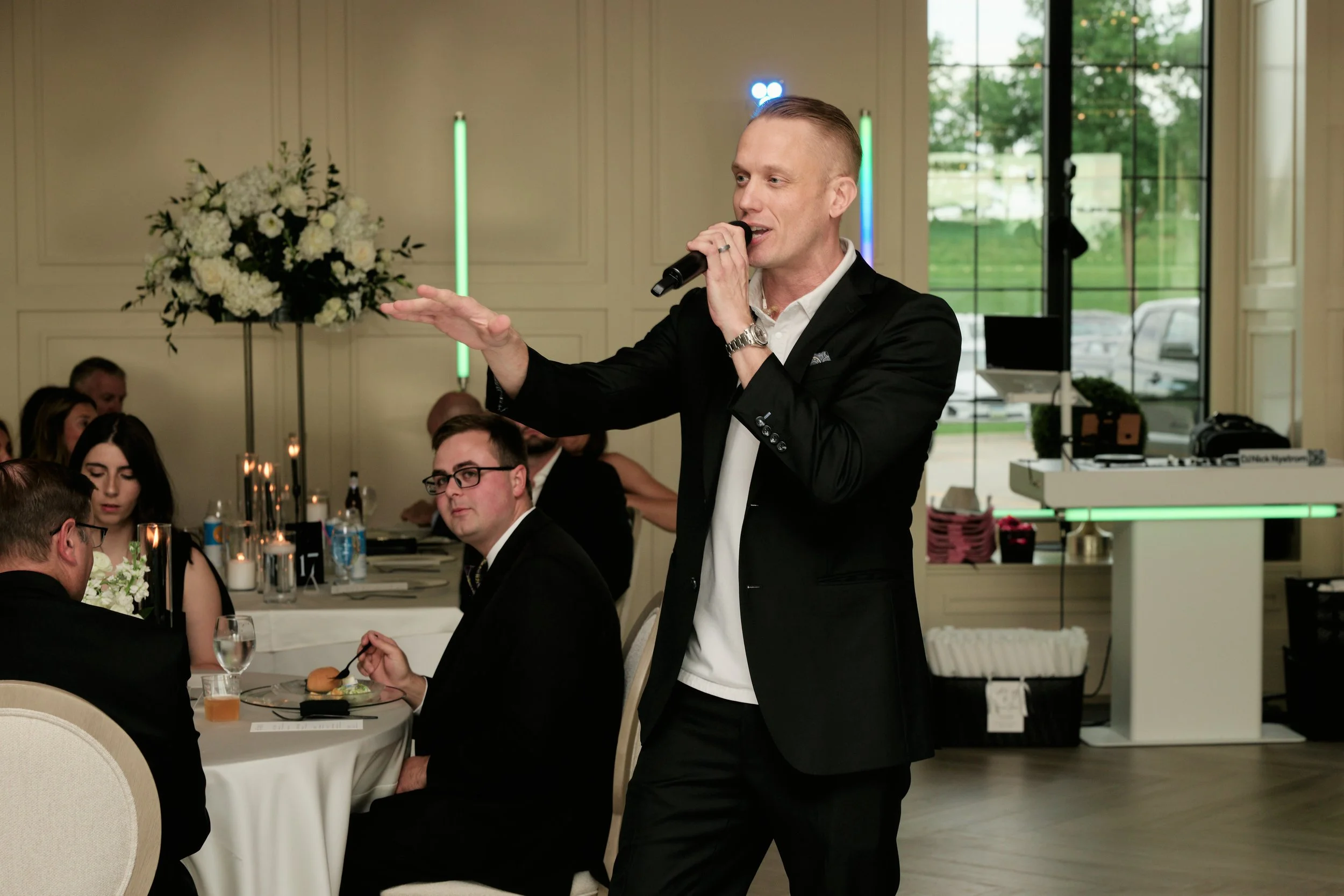 A man in a black suit is speaking into a microphone at a formal event with people seated at round tables inside a decorated room with large windows and floral arrangements.