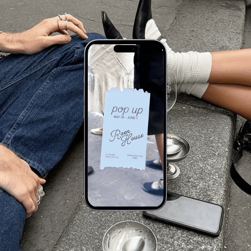 A person taking a photo of a receipt on their phone. The receipt reads "pop up, May 30 - June 1, Raen House." They are sitting on a bench, with a cup and other items around them, and another person's legs are visible in the background.