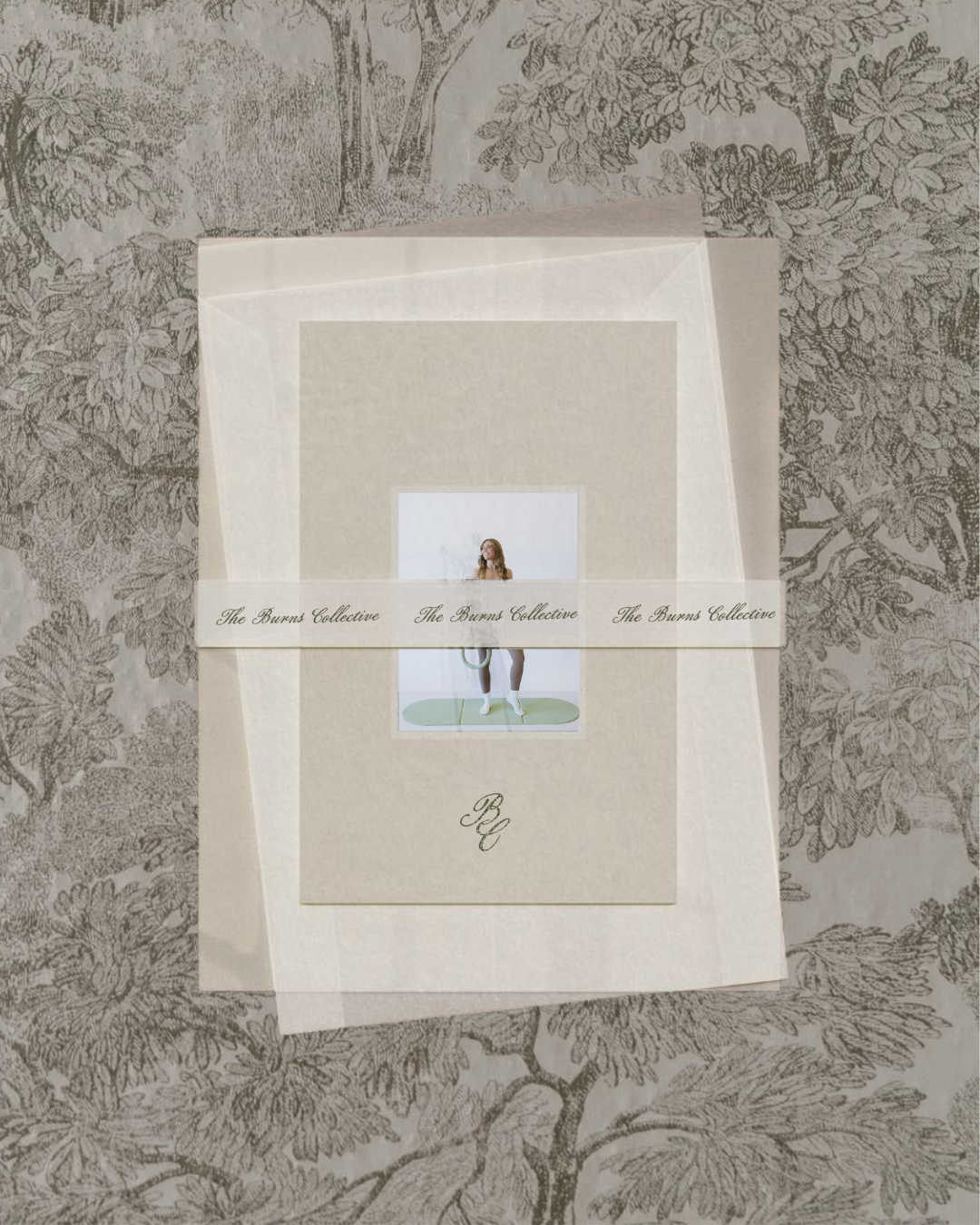 A paper invitation or card with a graphic of a woman exercising, tied with a white band that says 'The Burns Collective' repeatedly, resting on a printed tablecloth featuring tree and leaf illustrations.