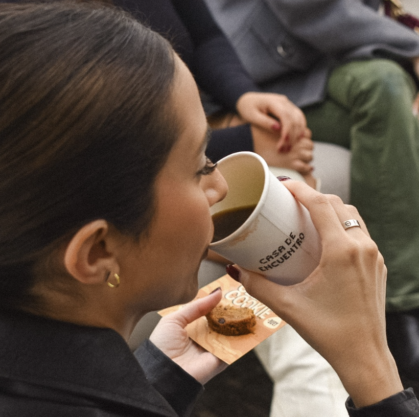 Woman drinking coffee from a white mug while holding a cookie in her other hand, with people sitting nearby.