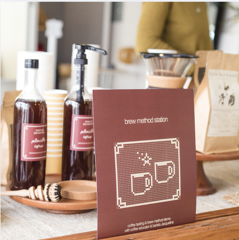 A coffee tasting station with bottles of flavored syrup, a sign titled 'brew method station' with an illustration of two coffee mugs, and various coffee supplies on a wooden table.