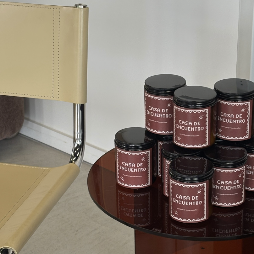 A round wooden table holding several black jars with maroon labels that read 'Casa de Encuentro'. Part of a beige chair with a chrome leg is visible to the left of the table.