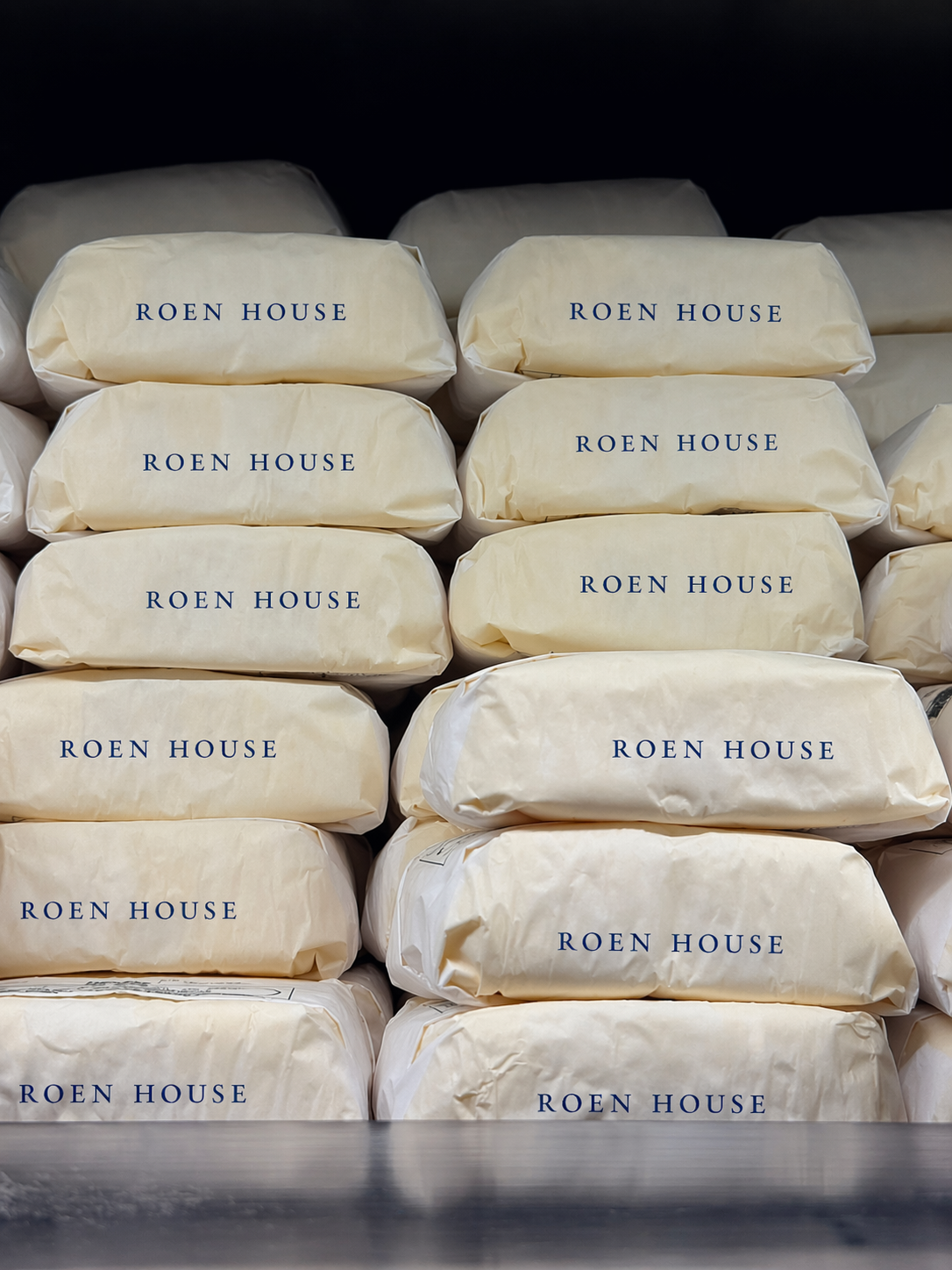 Stacked packages wrapped in cream paper with 'ROEN HOUSE' printed in blue, on a black background.
