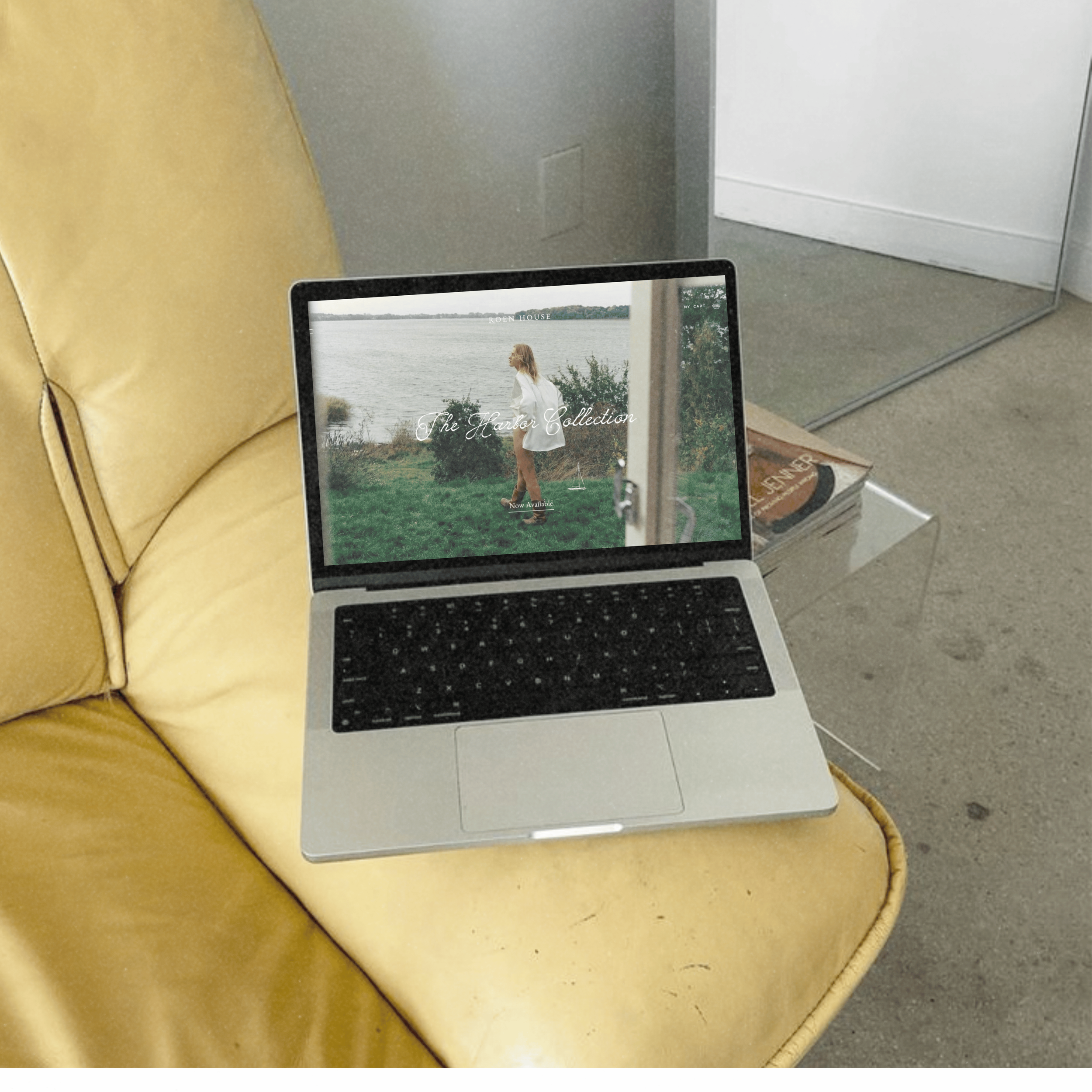 A laptop on a table displaying a website with an image of a woman by the water, with a yellow cushion and a white wardrobe or cabinet in the background.