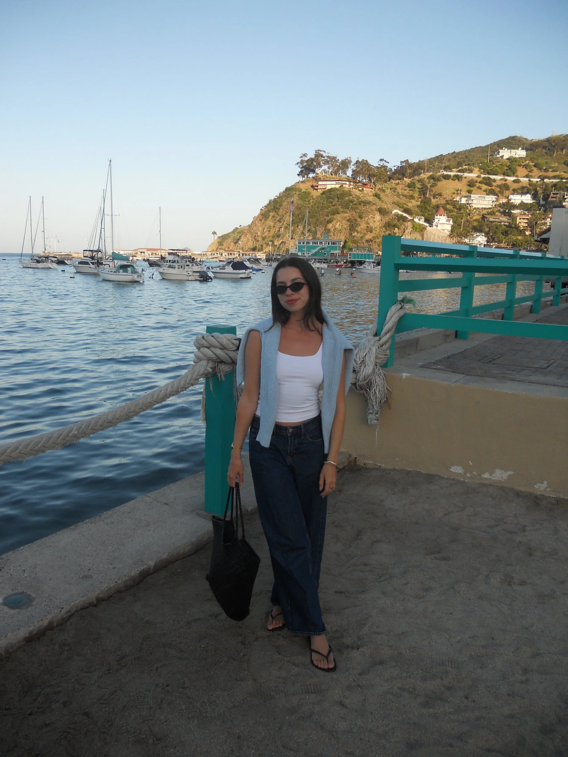 A woman with dark hair, wearing sunglasses, a white top, blue jeans, and flip-flops, standing on a concrete pier by a body of water with boats and a hillside in the background.