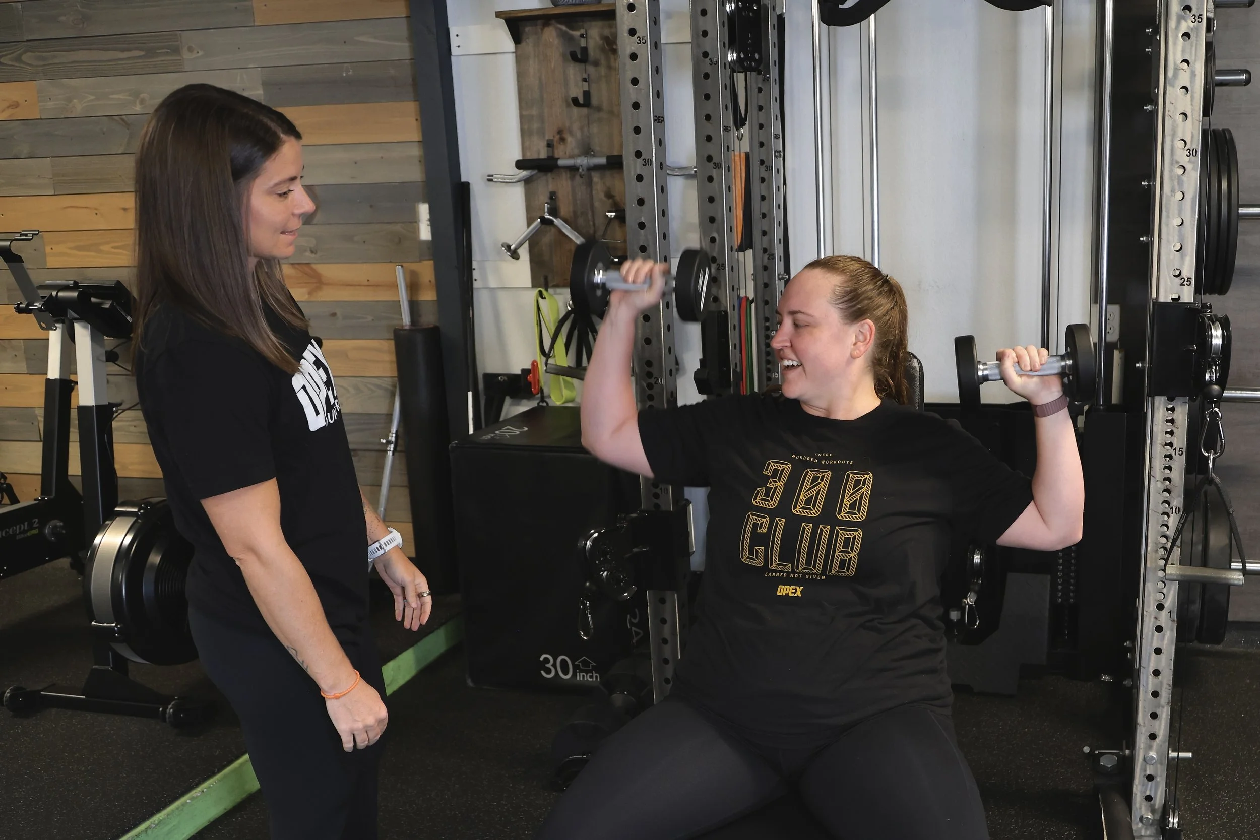 A woman is spotting another woman performing a shoulder pres with a dumbbell in a gym, with gym equipment and wooden wall in the background.