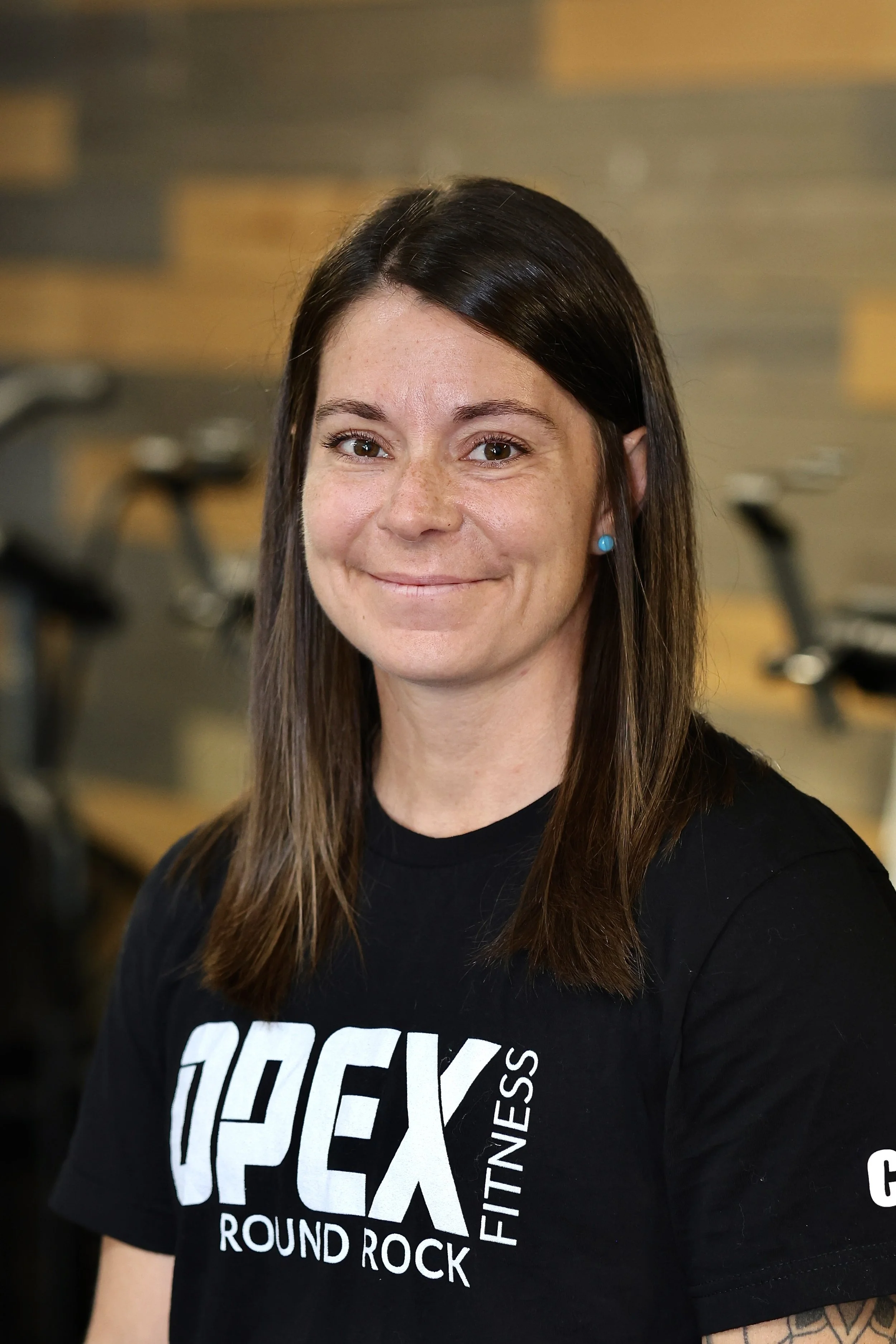A woman with long brown hair and blue earrings, smiling, wearing a black T-shirt with white text that reads 'ROCKY'. The background includes exercise bikes and a wooden wall.