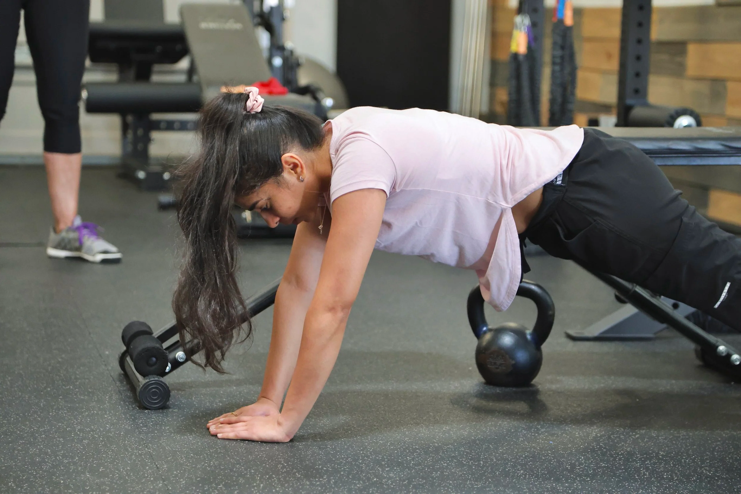 A woman in a pink t-shirt and black shorts performing a plank exercise on the floor of a gym, with her hands and toes touching the ground.