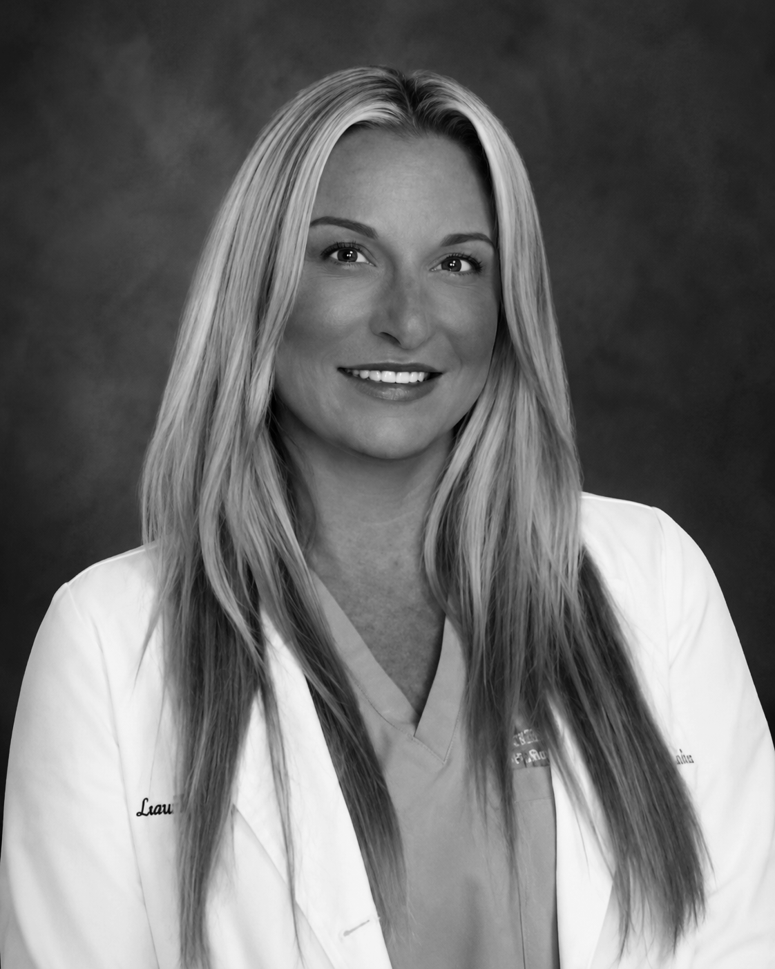 Black and white portrait of a smiling woman with long, straight hair wearing medical scrubs and a white coat.