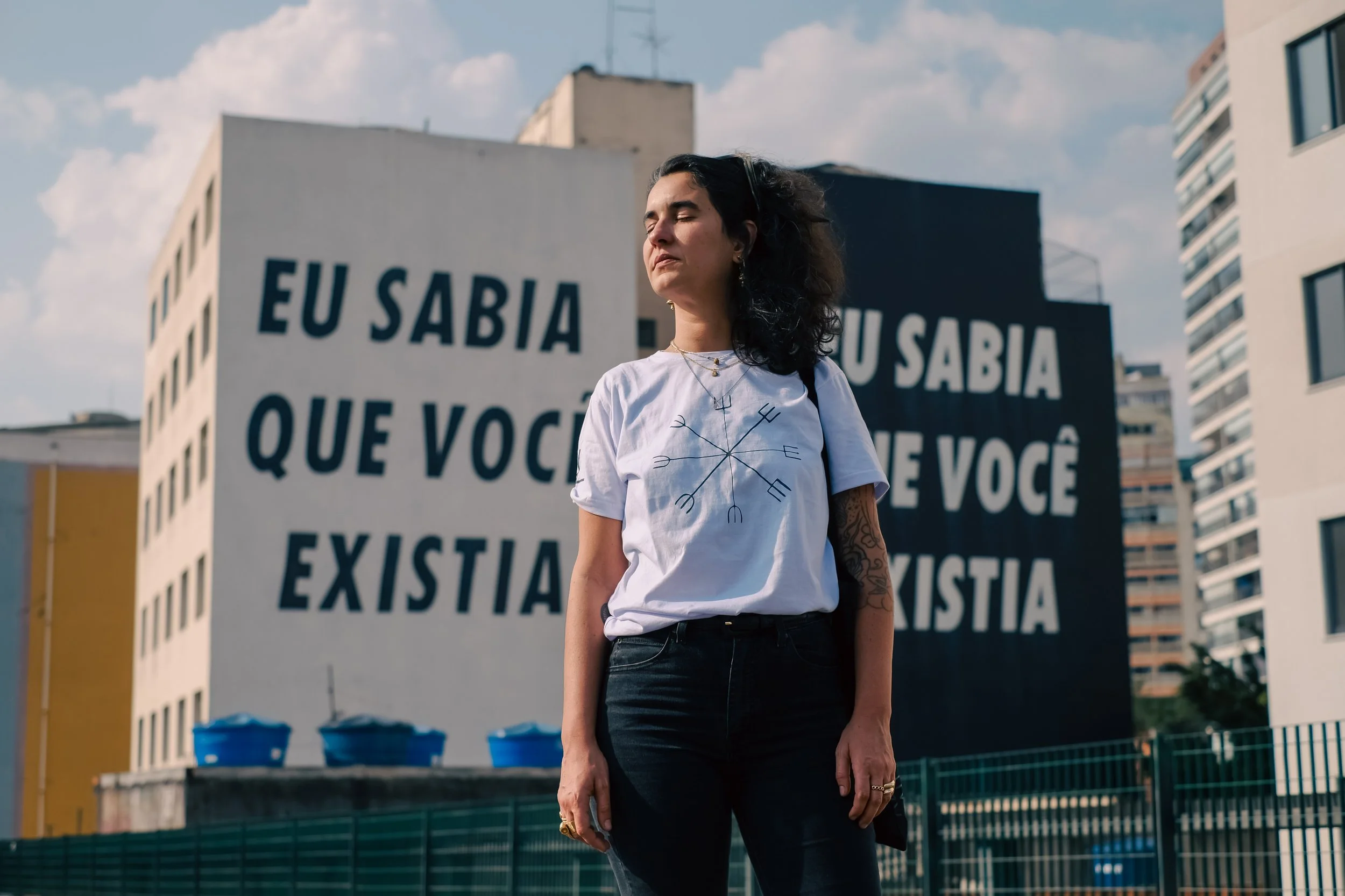 A woman with dark hair, closed eyes, and tattoos on her arm standing outdoors in front of a building with large black and white signs that say "EU SABIA QUE VOCÊ EXISTIA" in Portuguese.