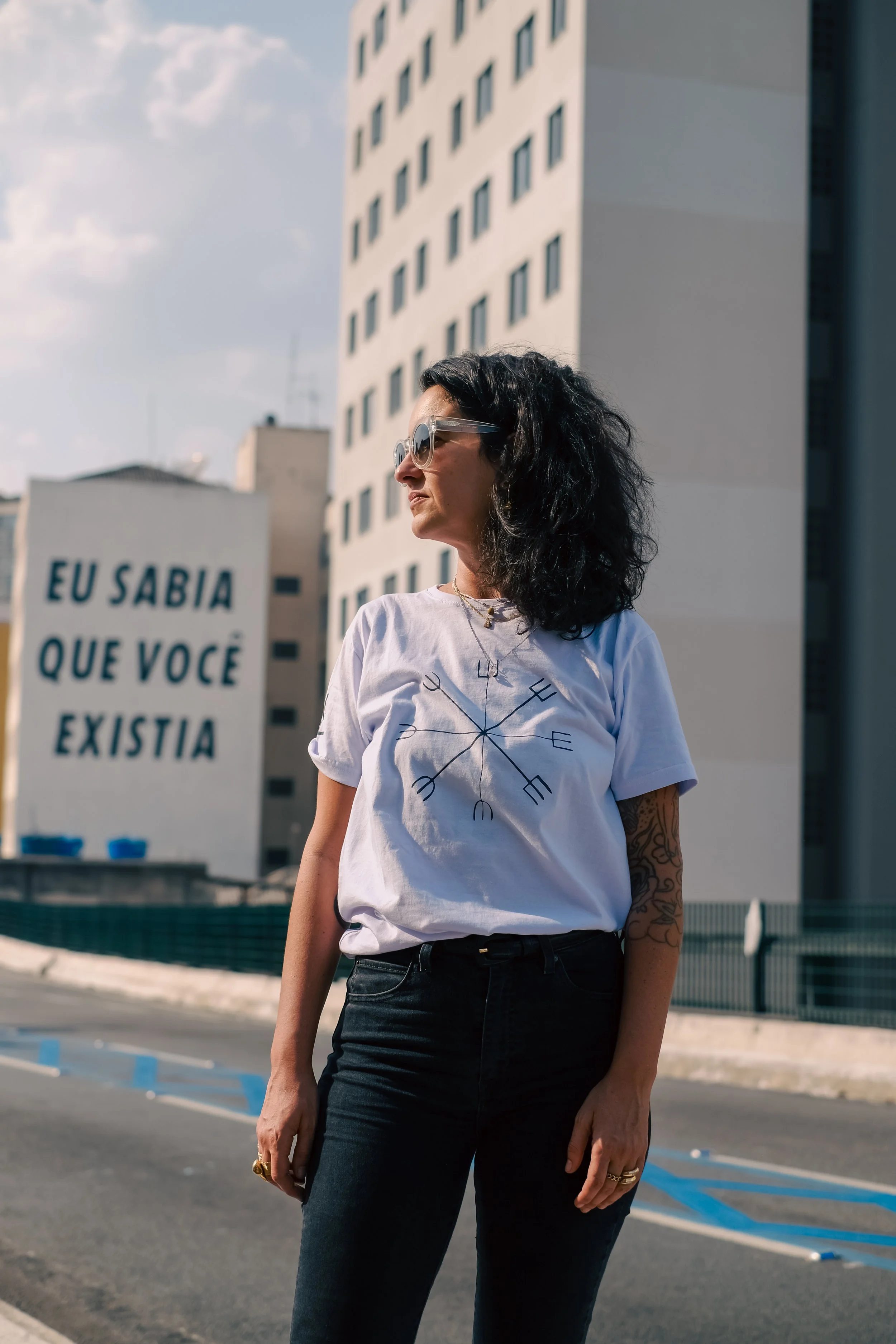 A woman with black, curly hair wearing sunglasses, a white t-shirt with a compass design, and black pants stands outdoors against a tall building. A sign in the background, in Portuguese, reads 'EU SABIA QUE VOCÊ EXISTIA,' which translates to 'I knew