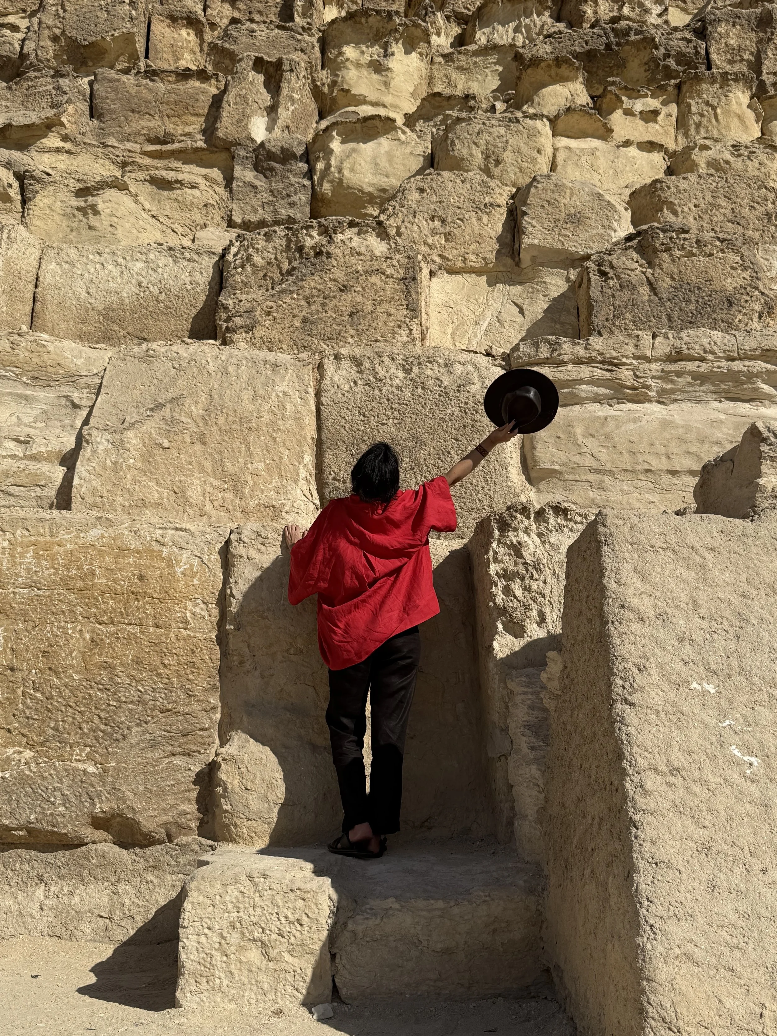A person wearing a red shirt and black pants reaching up with a black hat against a large ancient stone wall.