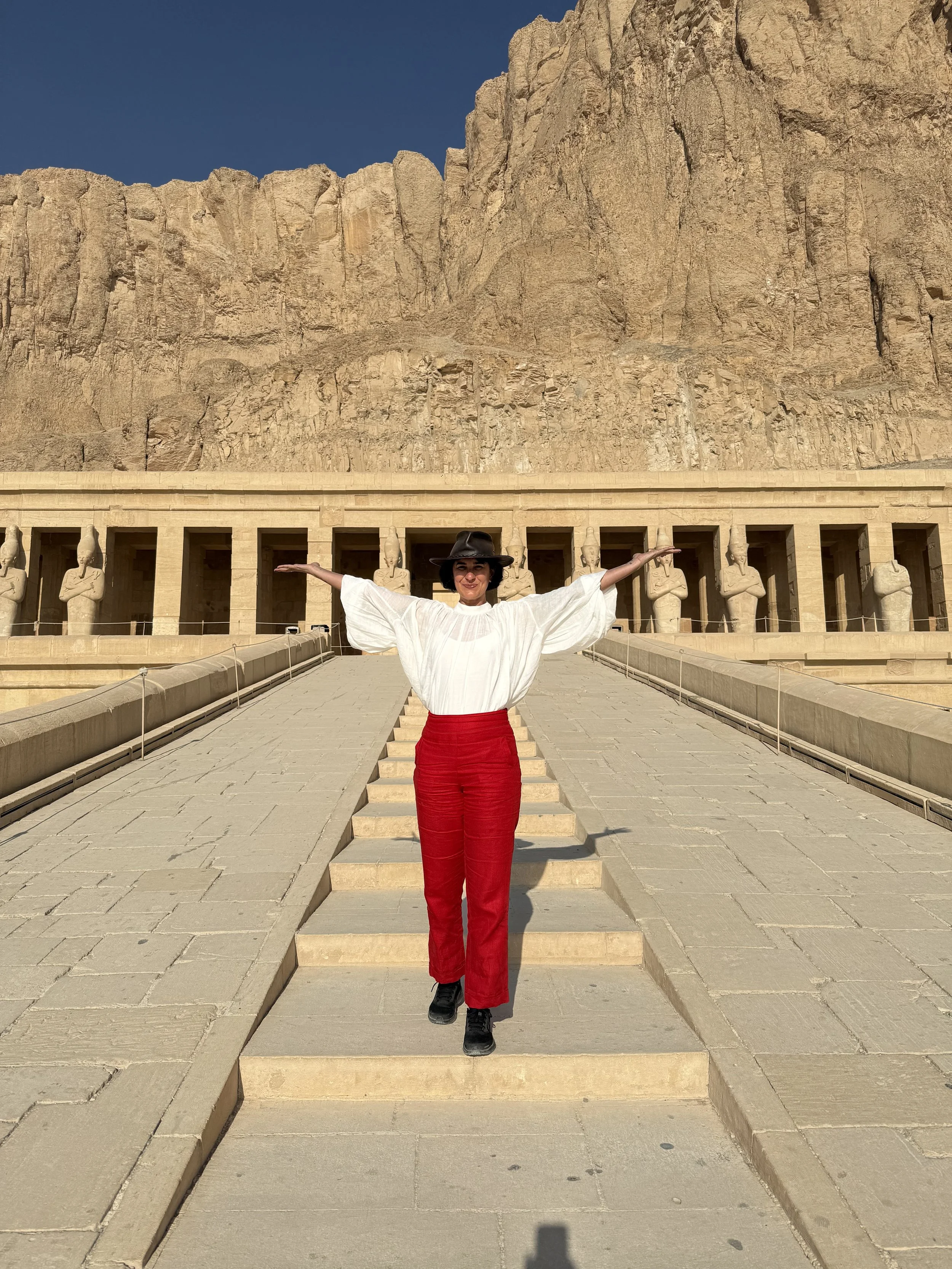A woman standing with arms outstretched on stone steps in front of ancient Egyptian statues and a large rock face at Luxor Temple in Egypt.