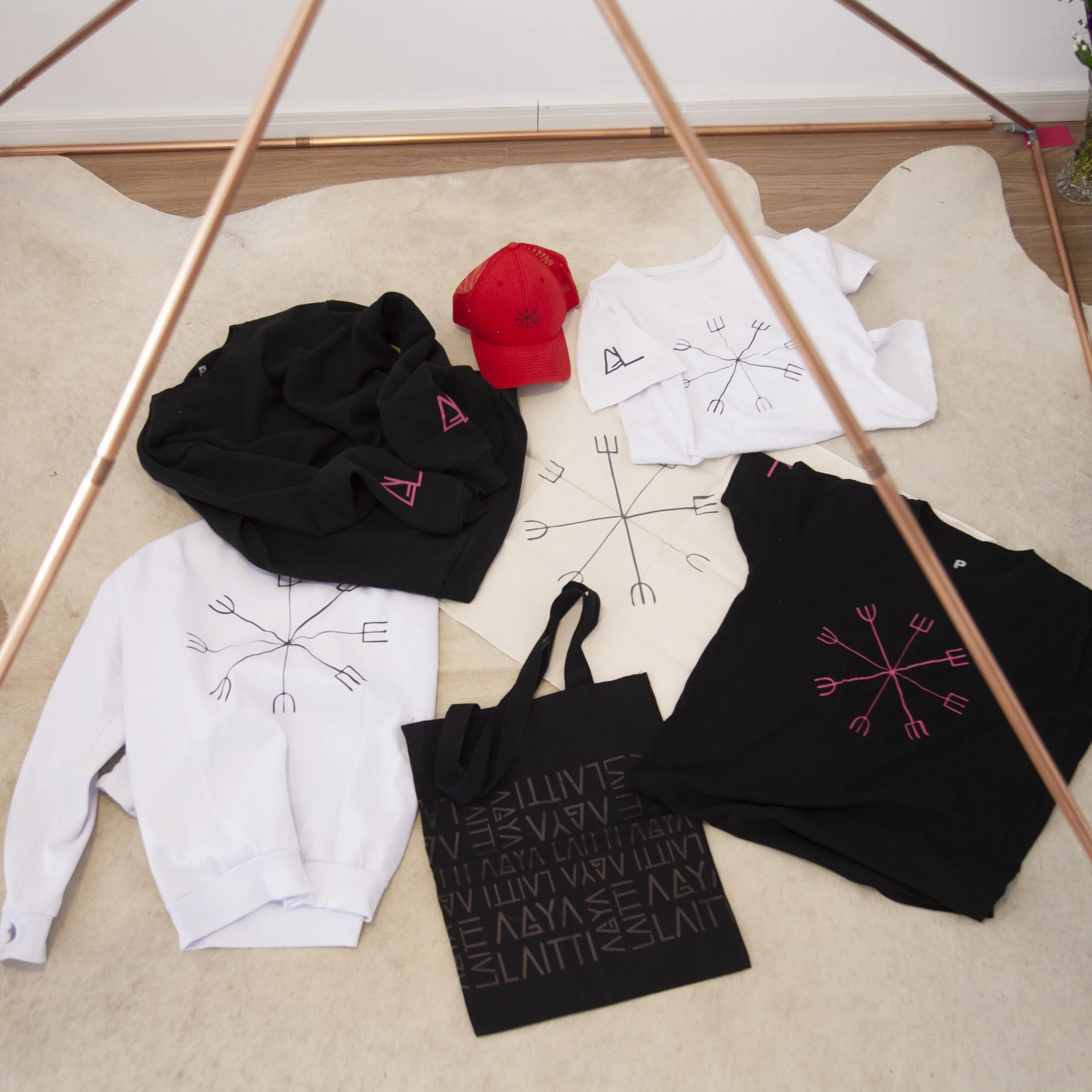 Clothing items with a black and pink logo, including a hoodie, sweatshirt, and T-shirts, laid out on a beige rug. A red baseball cap and a black tote bag with the same logo are also on the rug. Two white and two black T-shirts feature a pink and blac