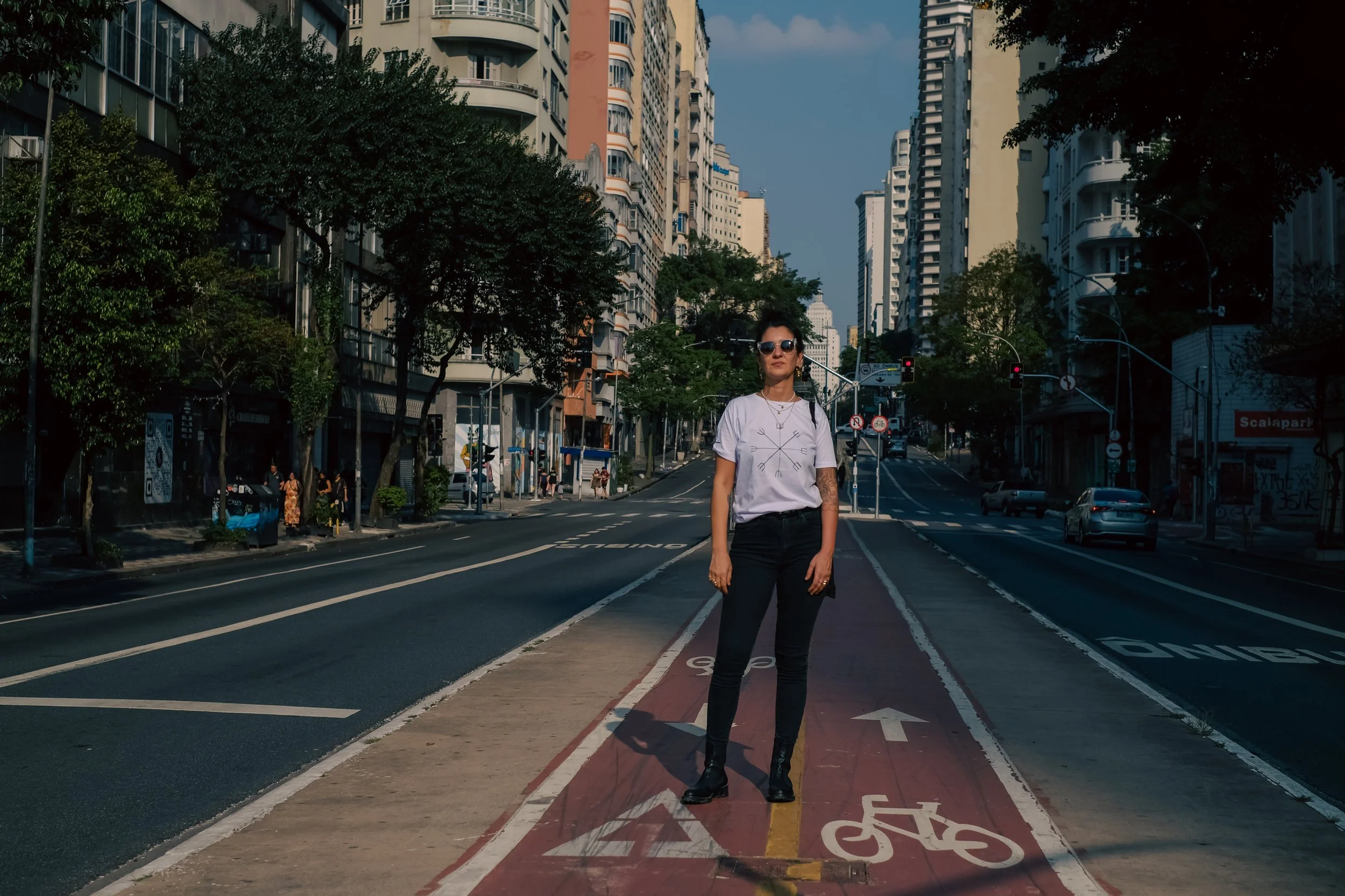 Me standing in the middle of a bike lane on a city street, with tall buildings and trees around her, wearing sunglasses, a white t-shirt, black pants, and black boots.