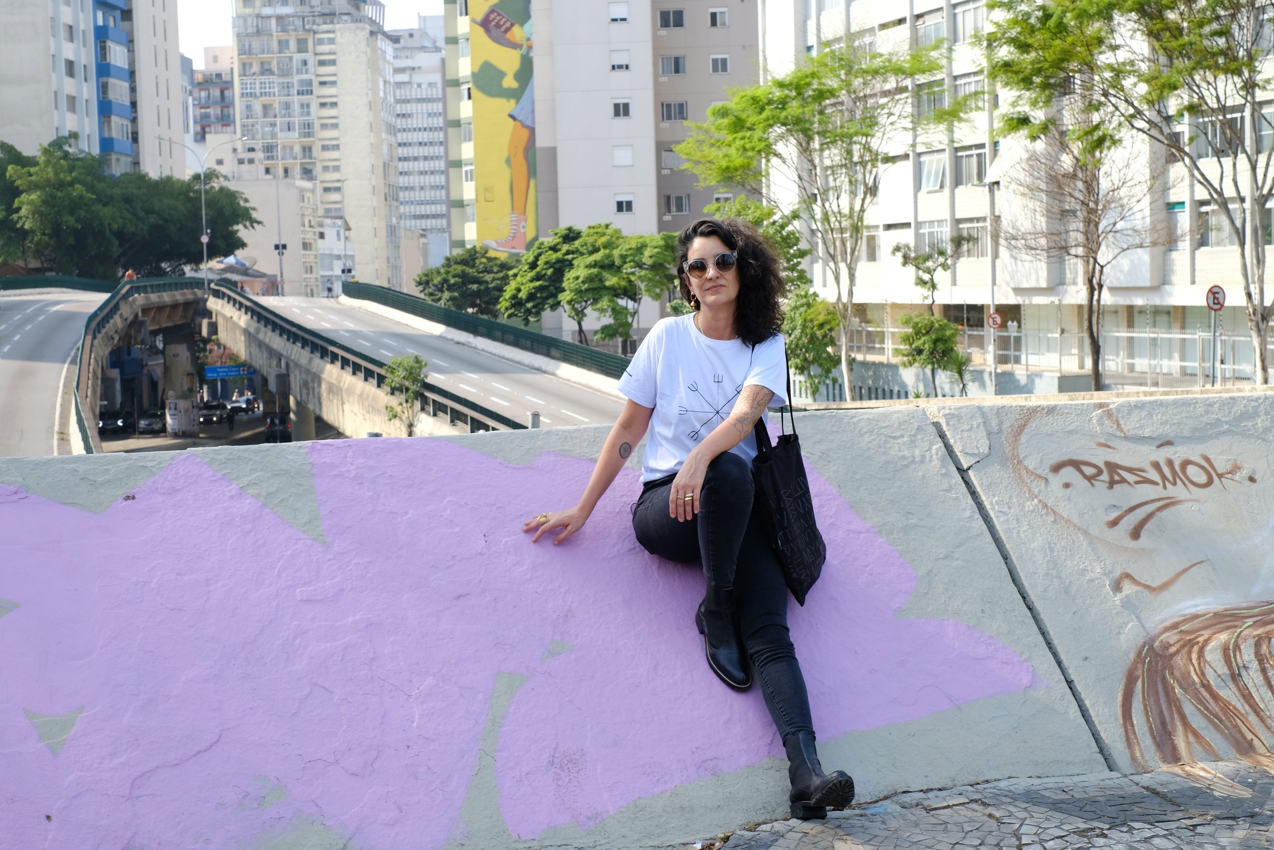 A woman with black curly hair, sunglasses, and tattoos is sitting on a colorful painted wall in an urban area with tall buildings and trees in the background.