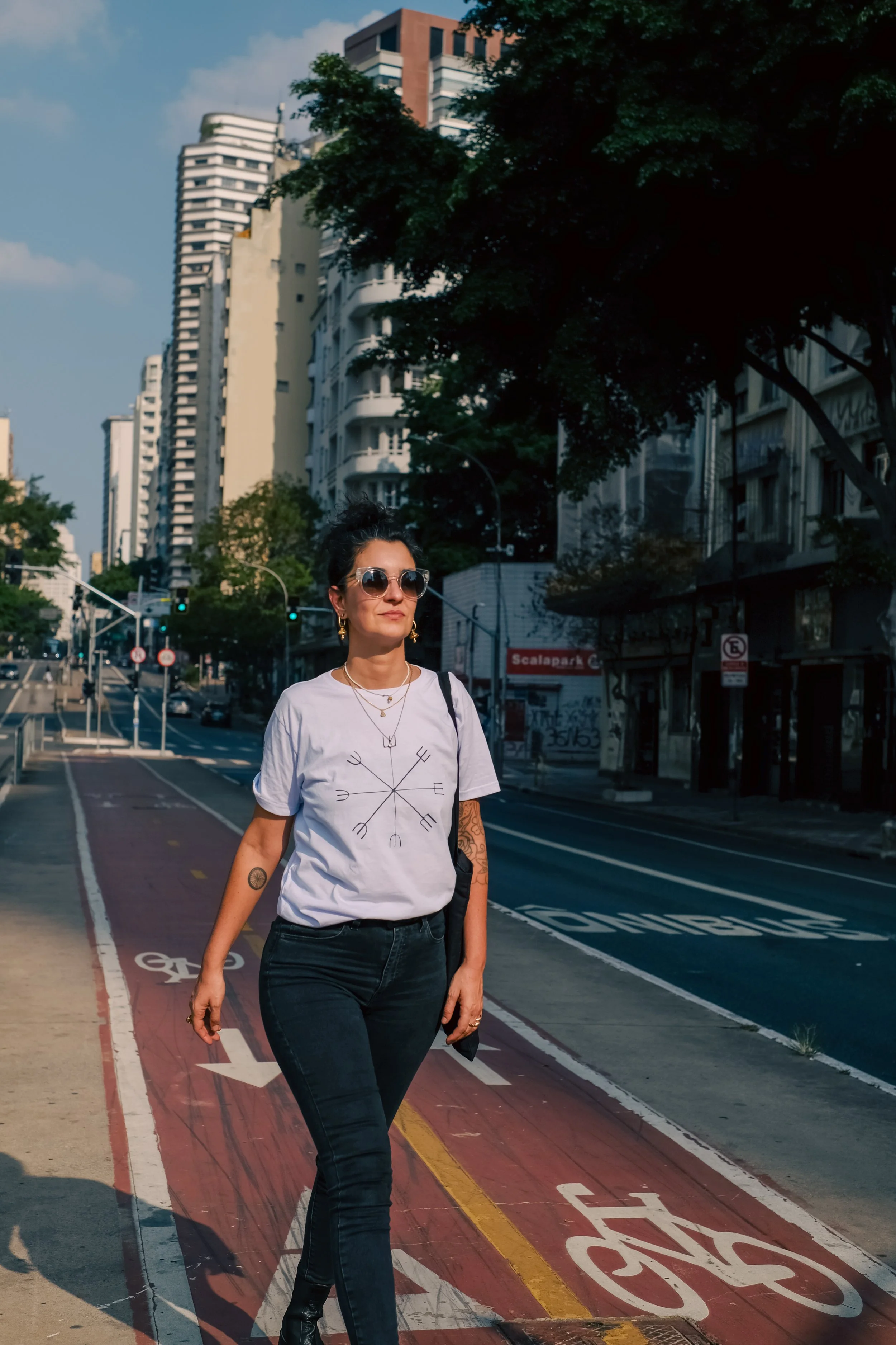 A woman walking on a city bike lane with high-rise buildings in the background. She is wearing sunglasses, a white t-shirt, dark jeans, and jewelry.