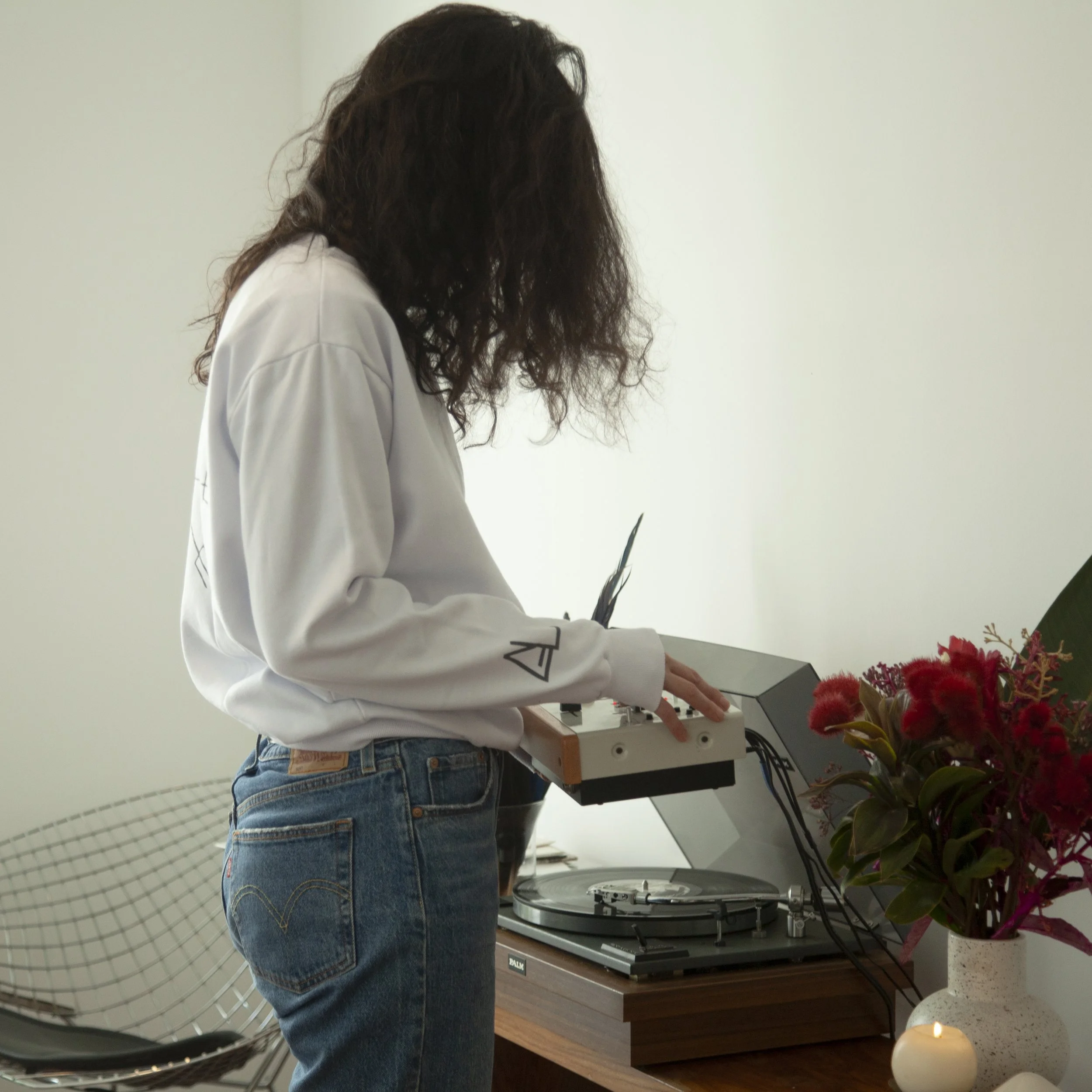 A person with curly hair wearing a white sweatshirt and blue jeans using a music synthesizer next to a turntable and a vase with red flowers and a lit candle.