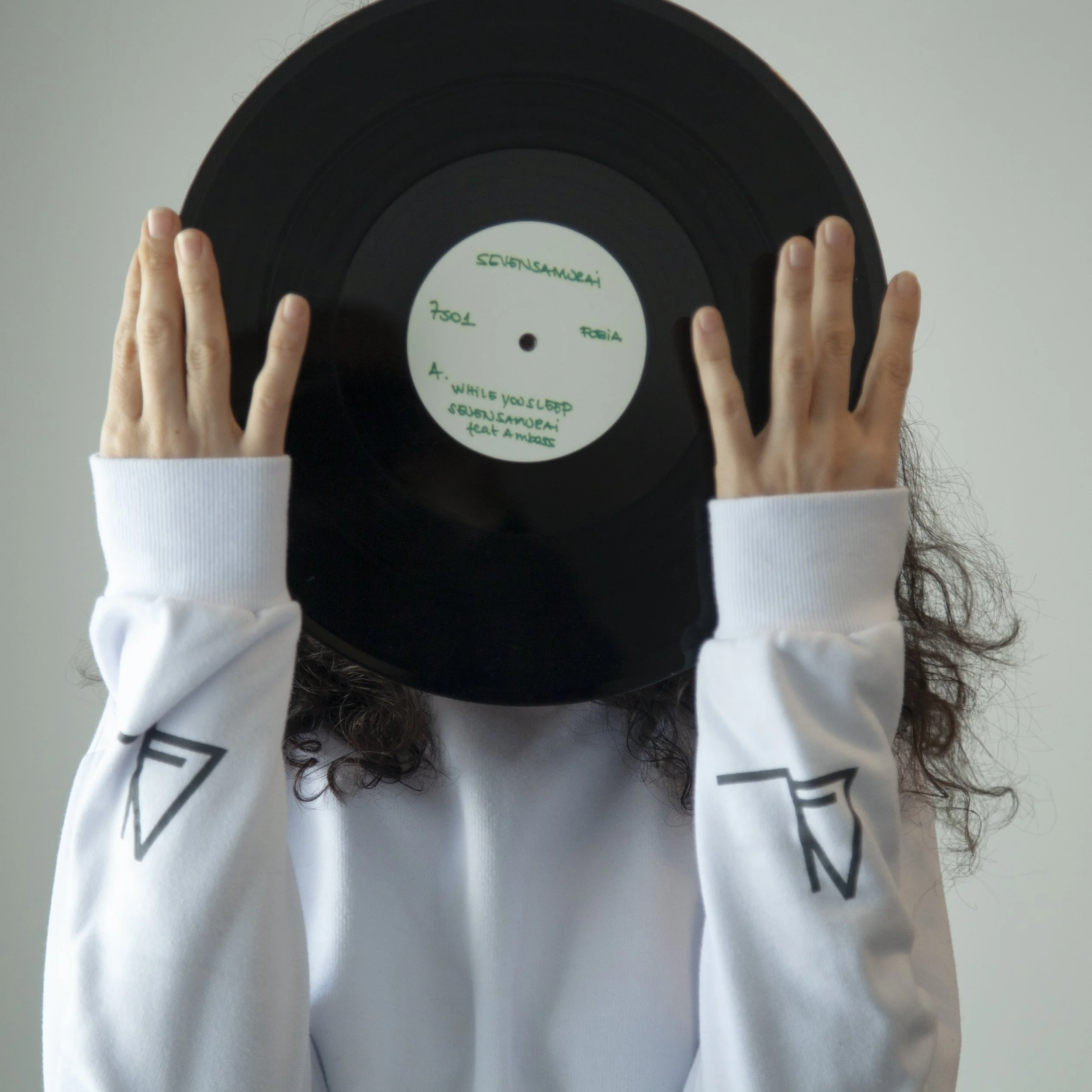 Person holding a black vinyl record in front of their face, with a white label that has green handwritten text, wearing a white long-sleeve shirt with black logos on the sleeves.