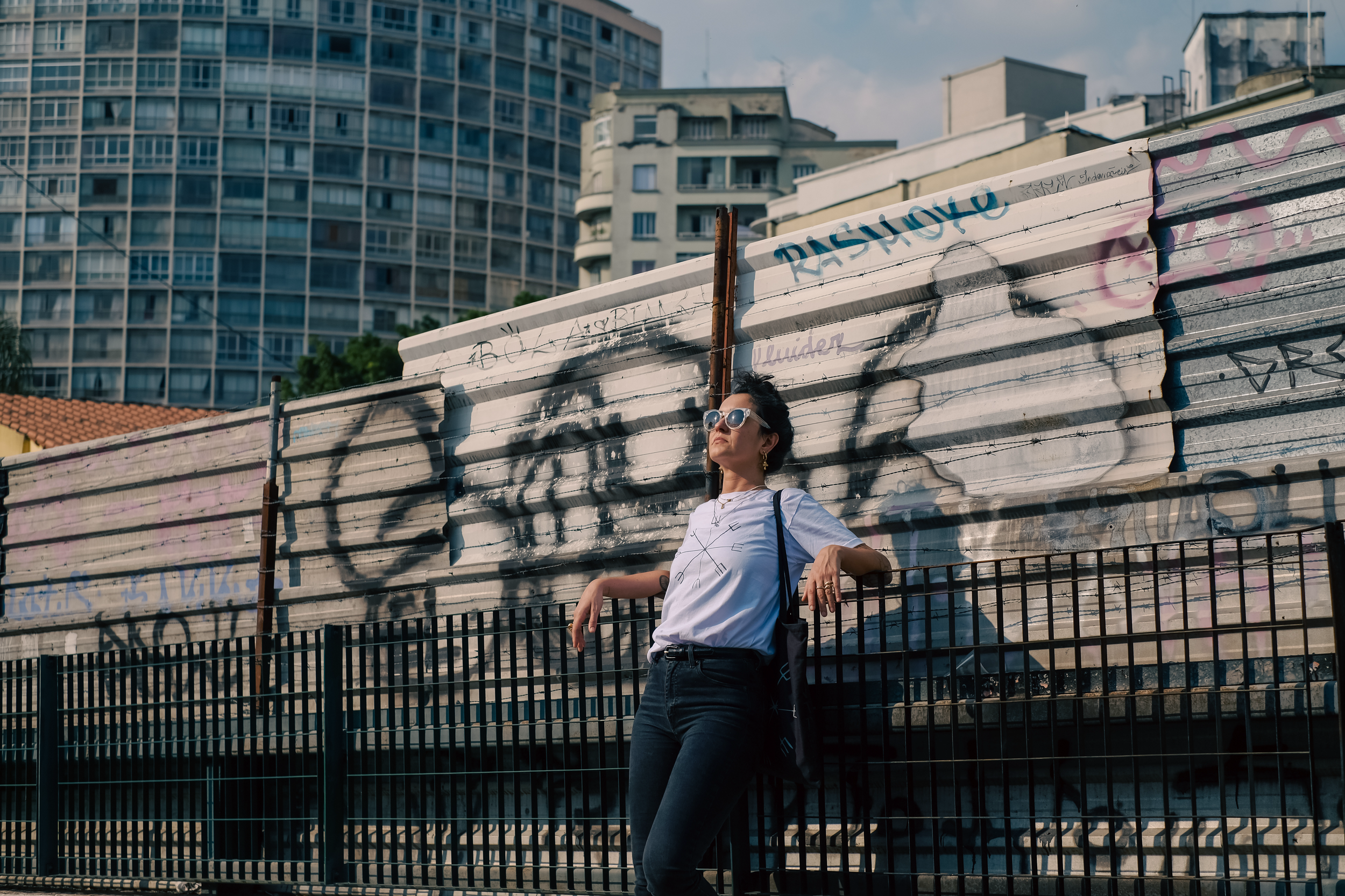 A woman wearing sunglasses and casual clothing leans against a black fence in front of a graffiti-covered metal wall, with modern high-rise buildings in the background.