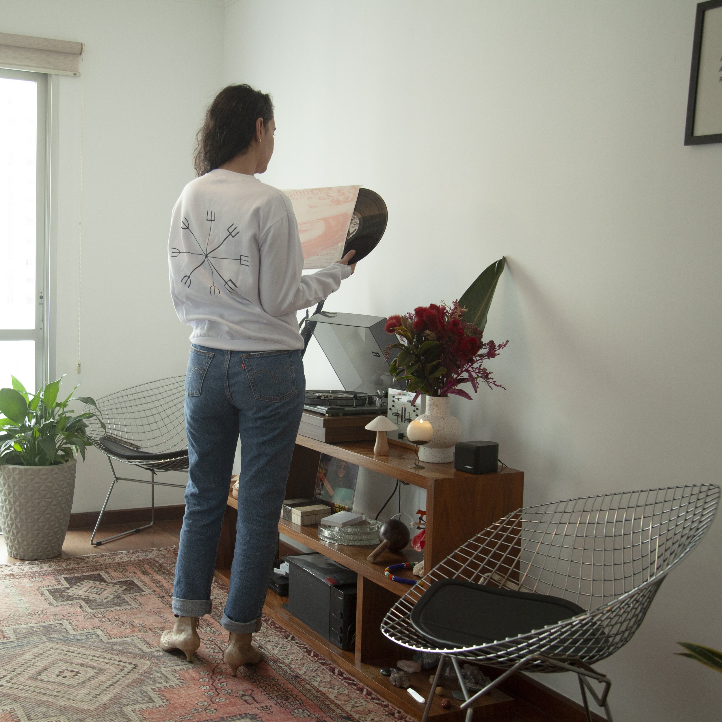 Woman with dark hair wearing a white sweatshirt and jeans, holding a vinyl record and sleeve, standing next to a wooden shelf with a record player, a large vase with flowers, and decorative items in a modern living room.
