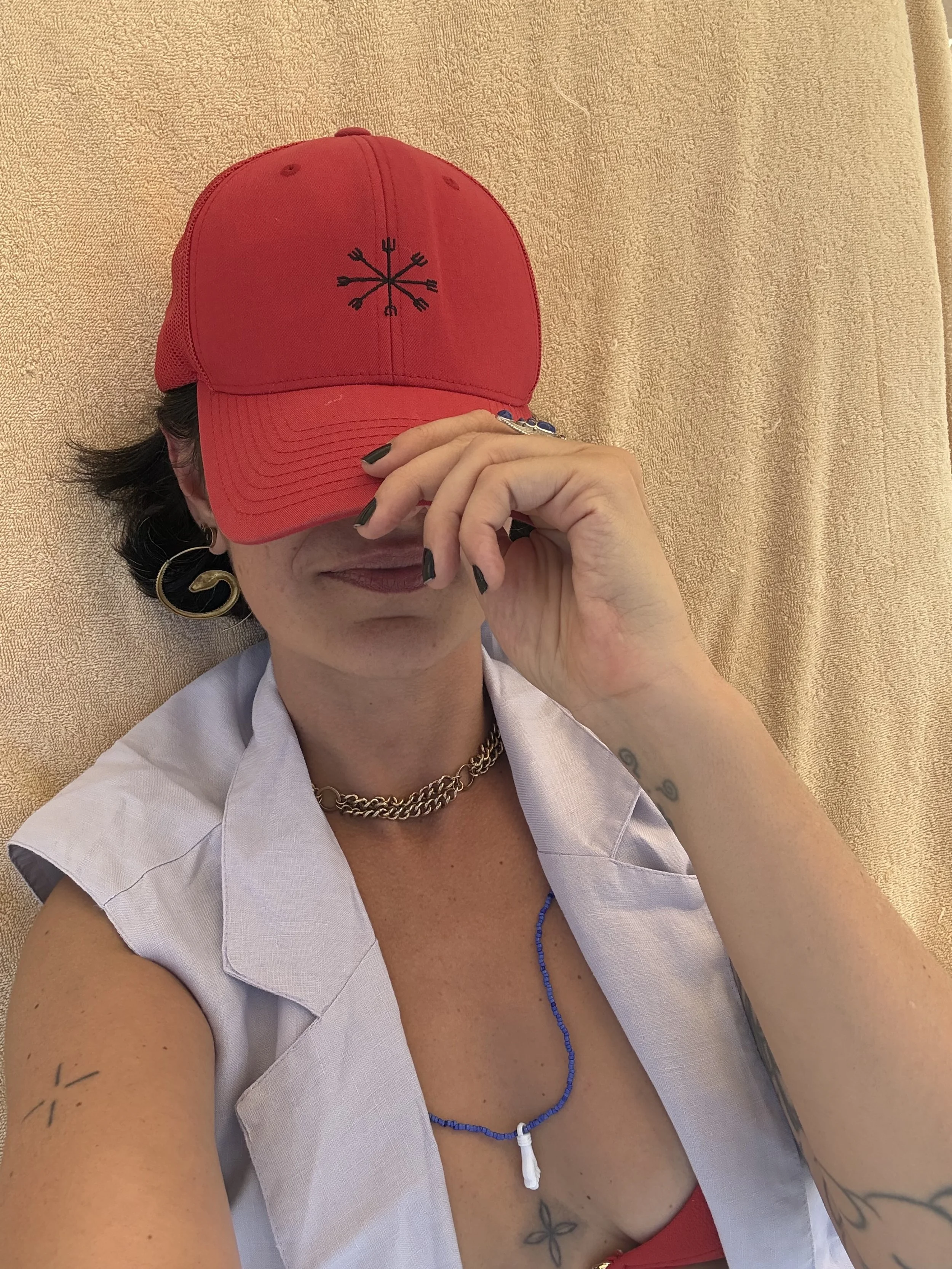 Woman lying down wearing a red baseball cap with a black symbol, white sleeveless shirt, gold chain necklace, blue beaded necklace, hoop earrings, and tattoos on arms and chest, holding the cap's brim over her eyes.