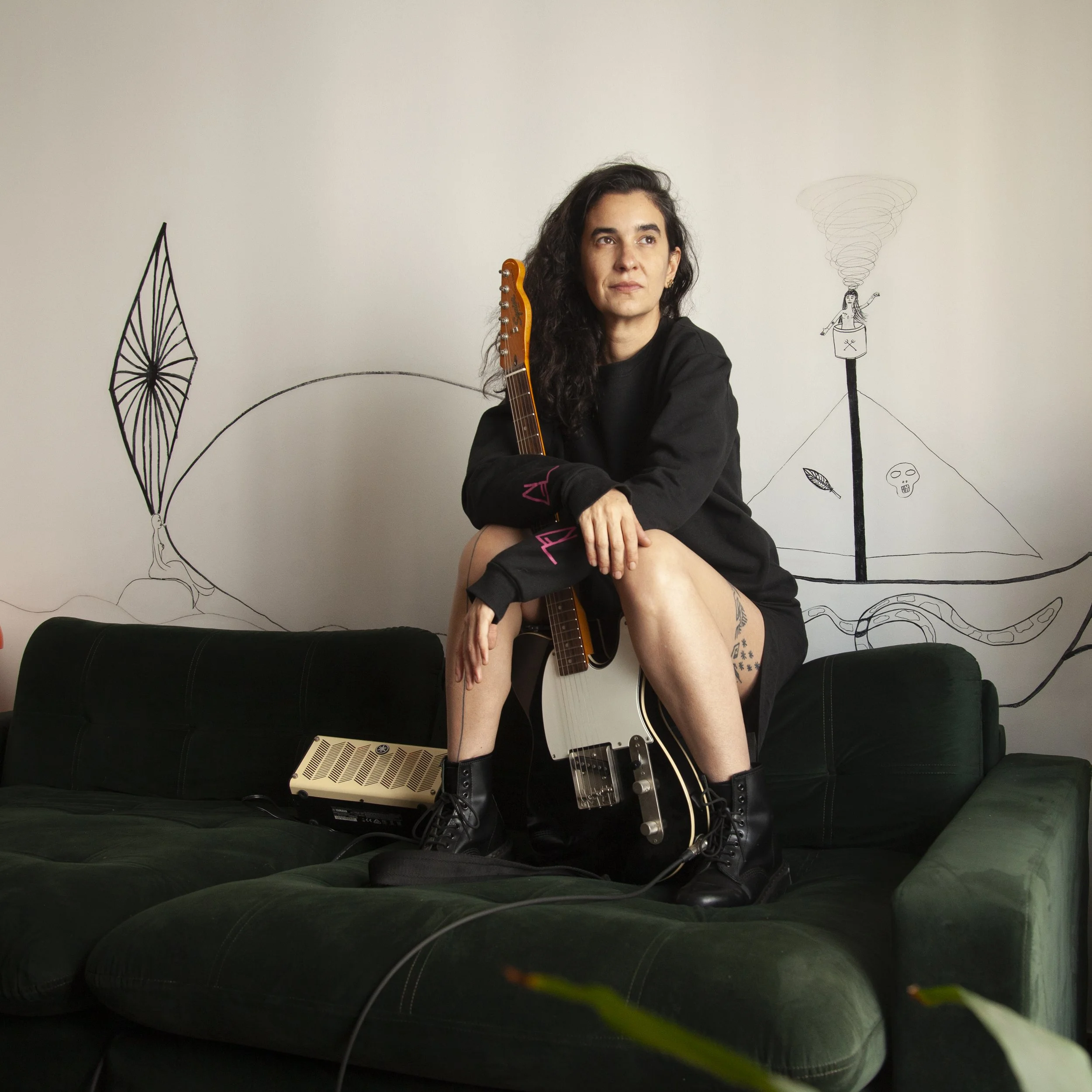 Woman with long dark hair sitting on a dark green couch, holding an electric guitar, wearing black boots and a black sweat-shirt, with drawings on the wall behind her.