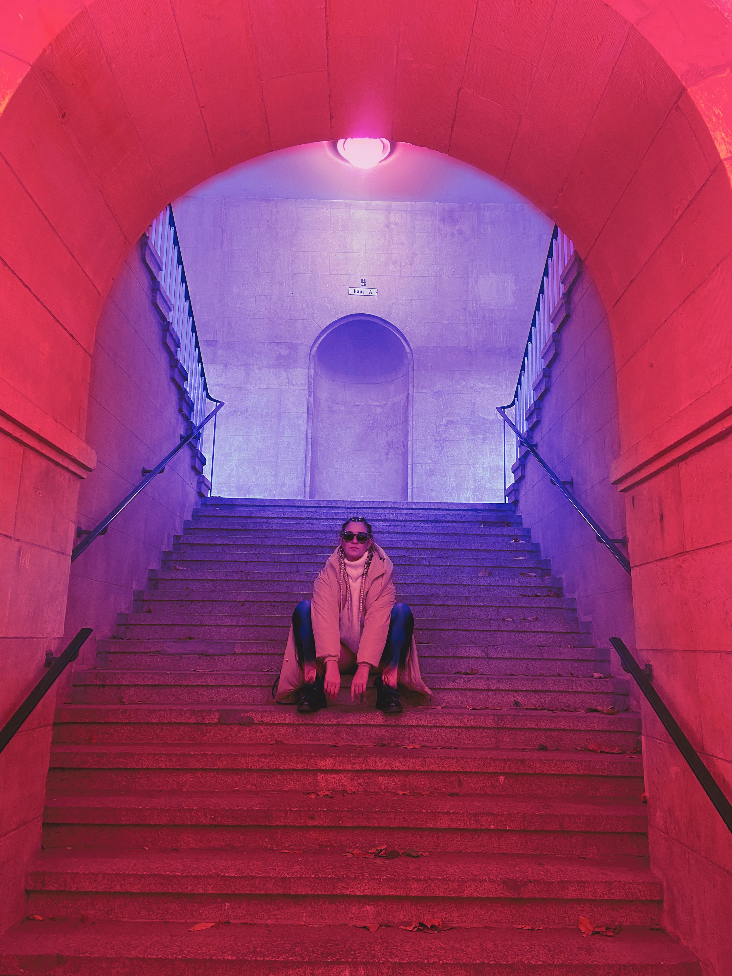 A woman sitting on stairs under a tunnel with vibrant red and purple lighting.