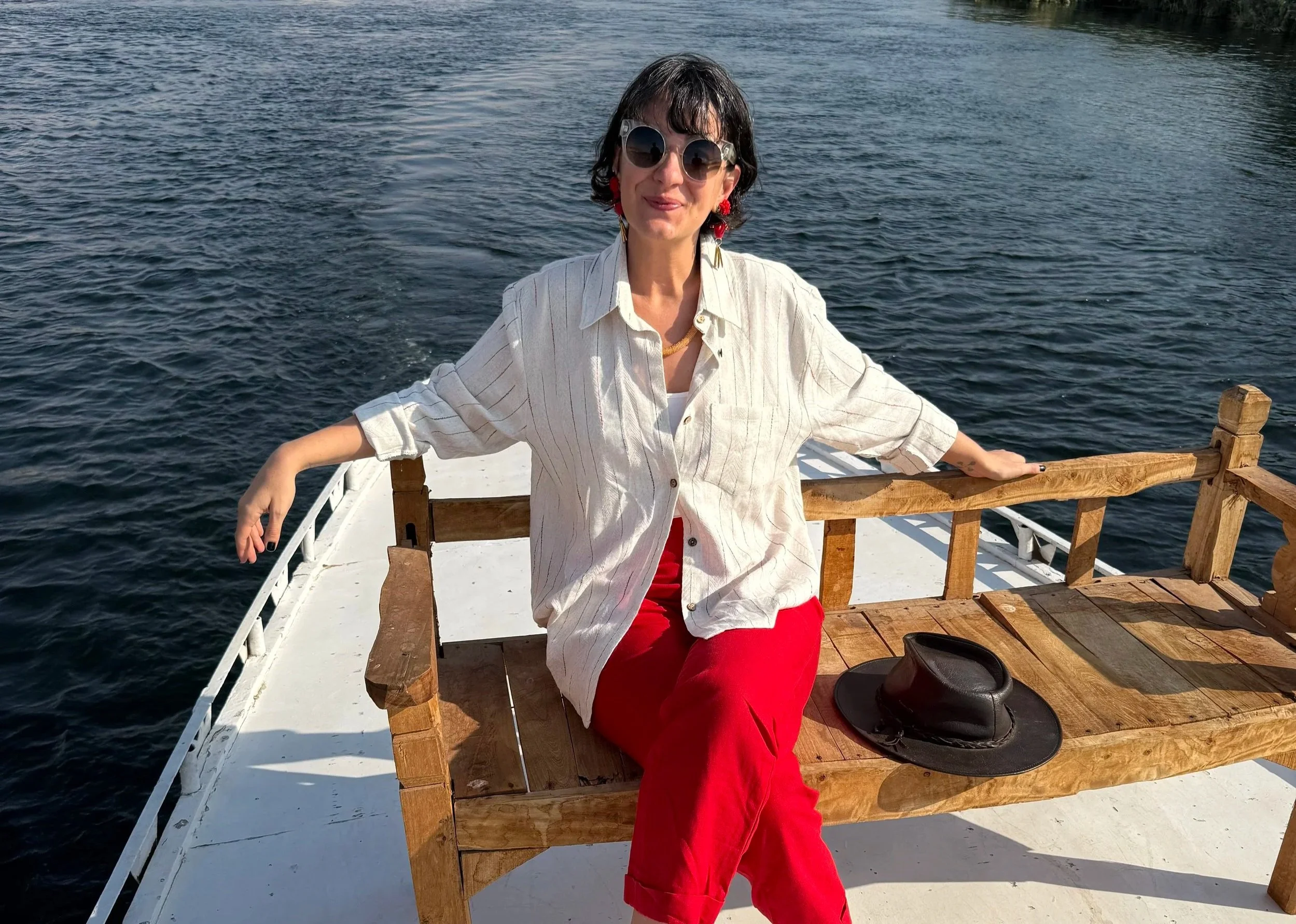 A woman with short dark hair and sunglasses sitting on a wooden bench on a boat, with water in the background. She is wearing a white shirt and red pants, and has a black hat placed on the bench beside her.