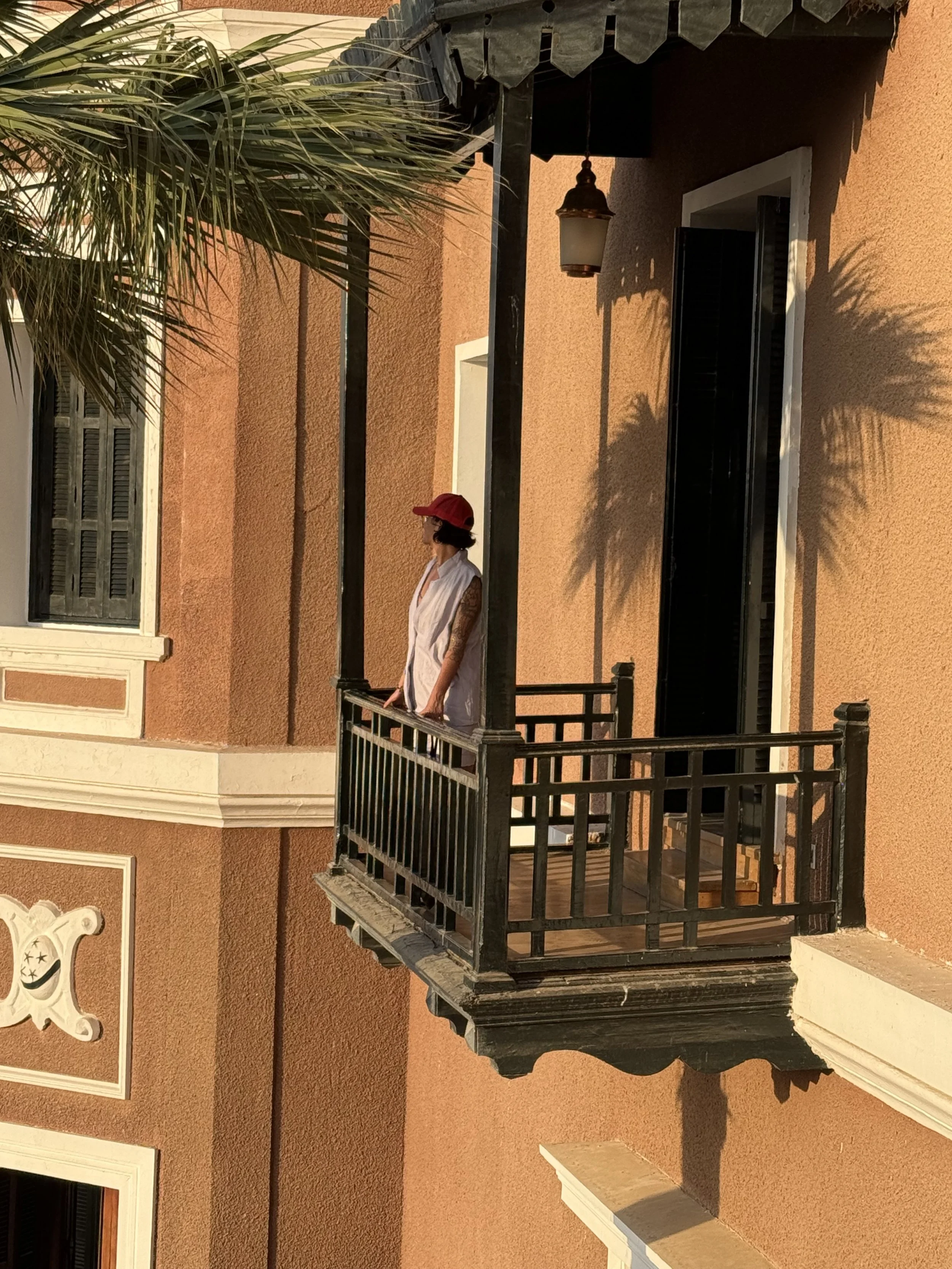 Woman standing on a small balcony of a peach-colored building, wearing a white top and red cap, with palm tree shadows on the wall.