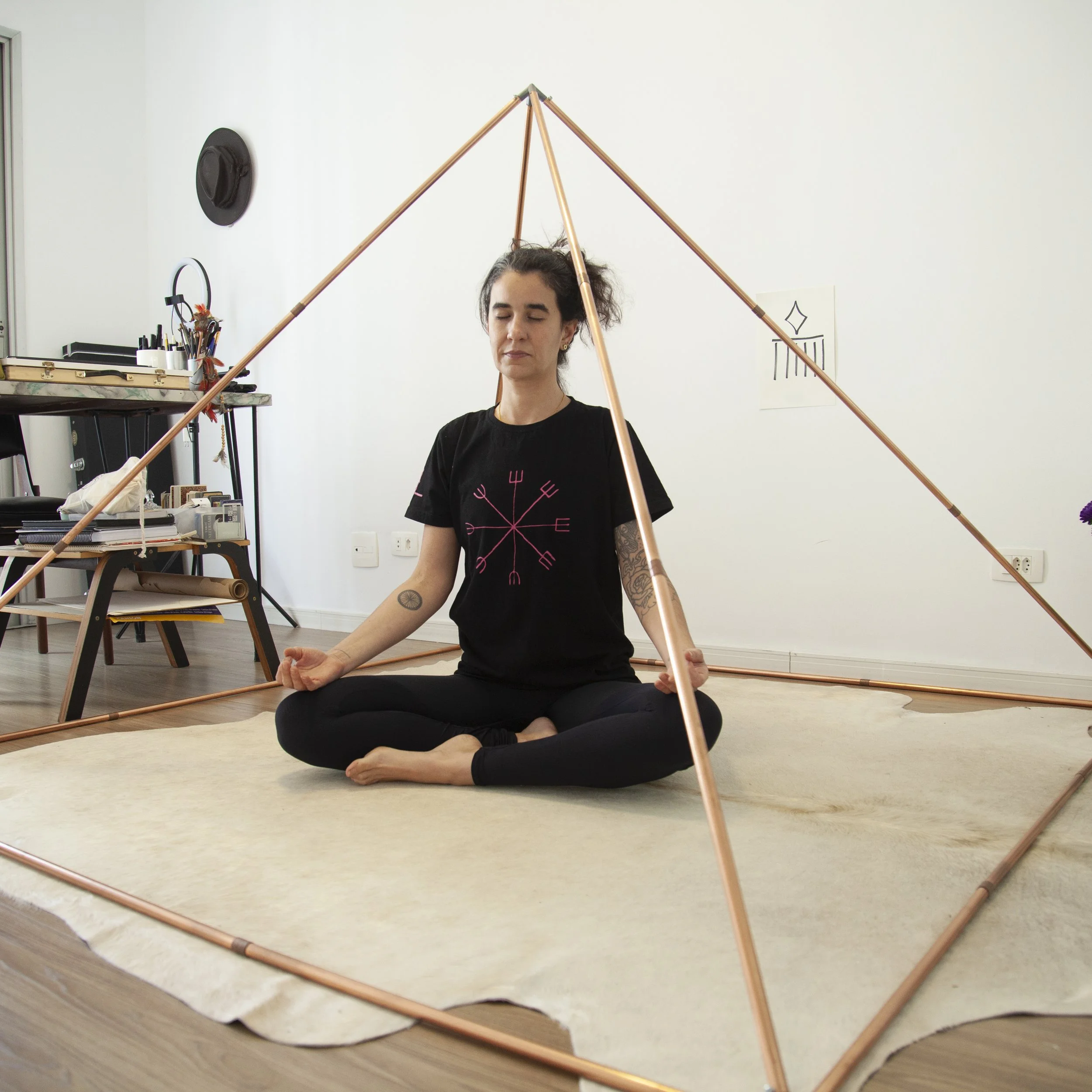 Woman meditating in a seated yoga pose inside a copper pyramid structure in a room with white walls and wooden floor.