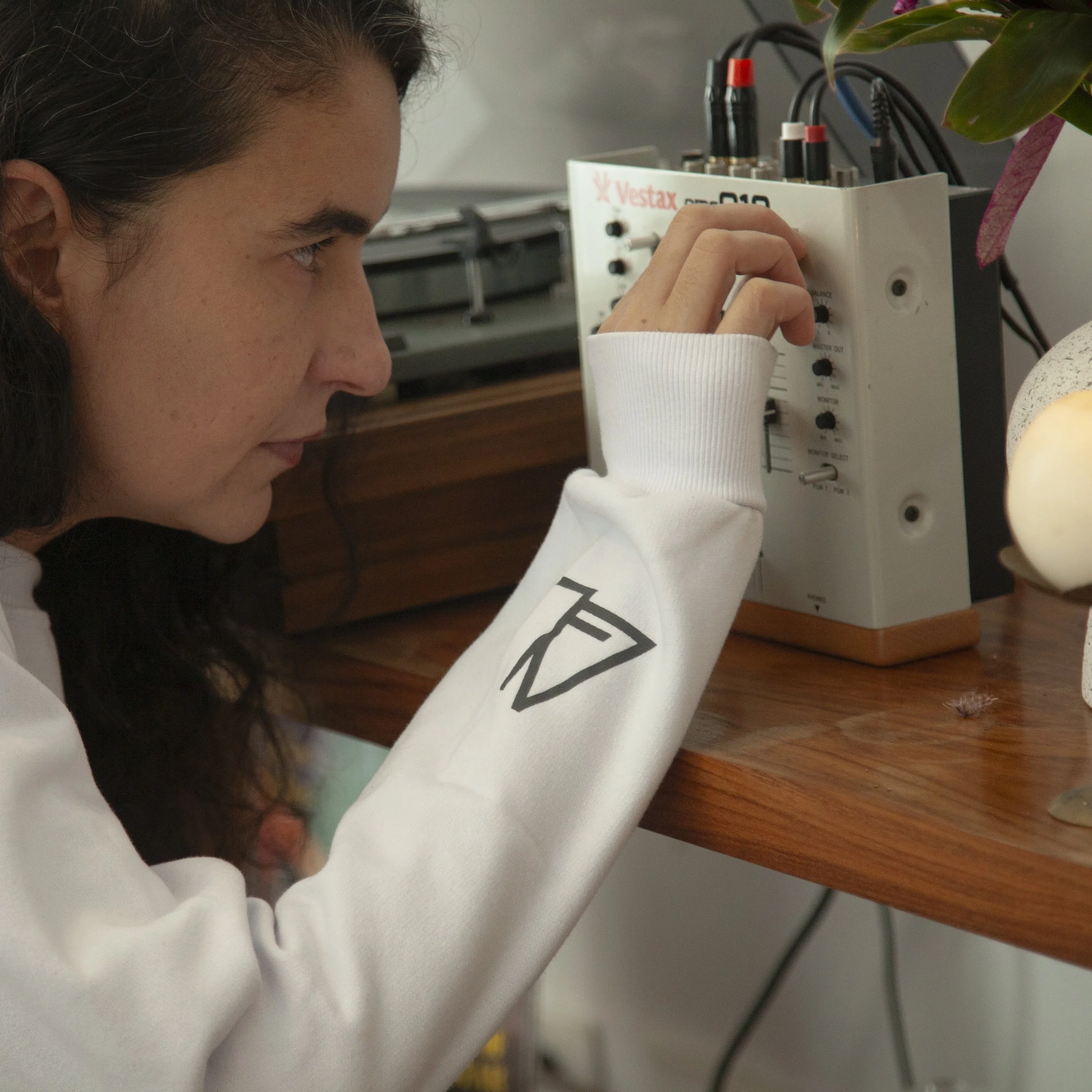 A young woman with dark hair working on an electronic device at a wooden workbench, adjusting controls on a white VESTAX audio mixer, with a potted plant nearby.