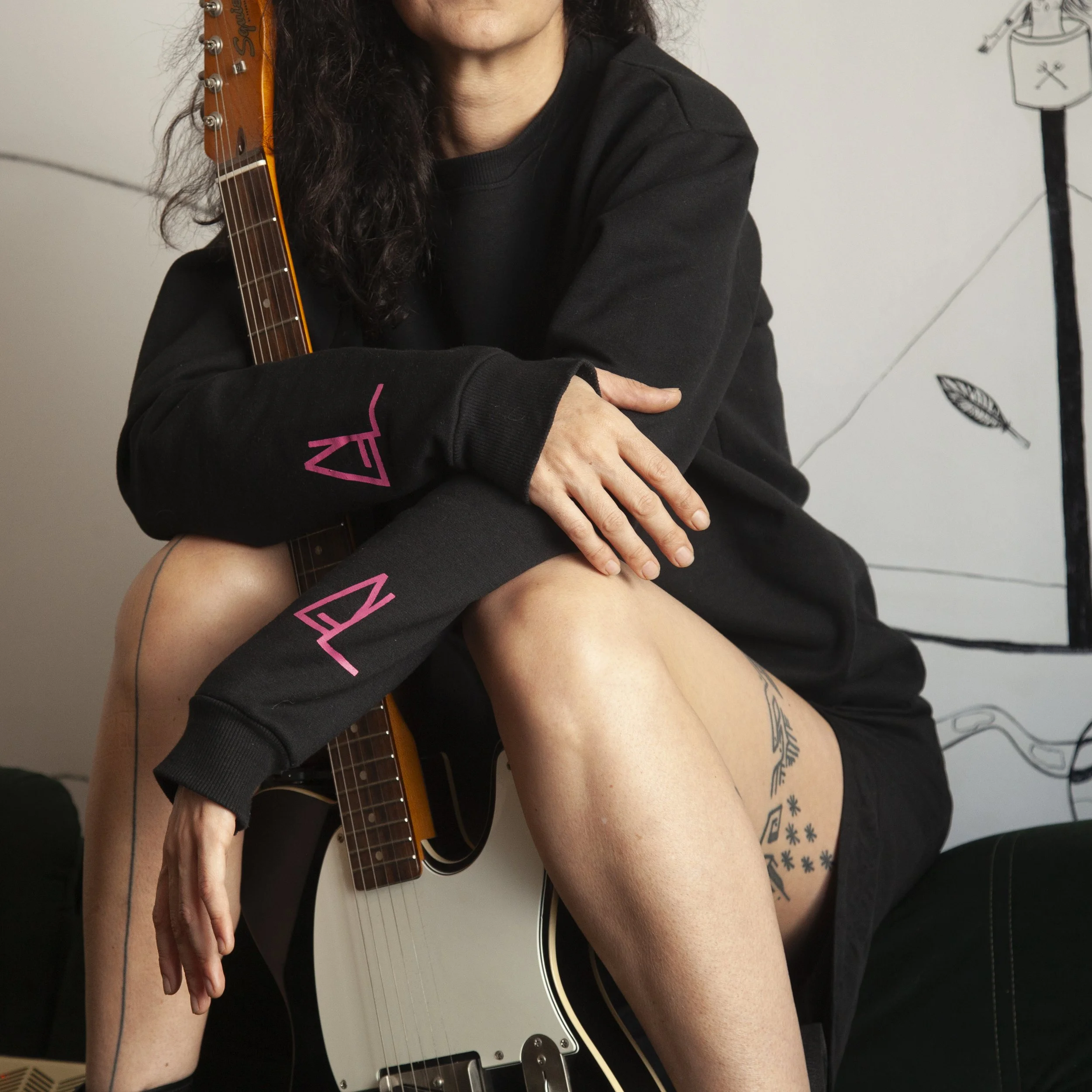 A person sitting with a guitar, wearing a black hoodie with pink lettering, and displaying tattoos on their legs. The person is in a room with wall art in the background.
