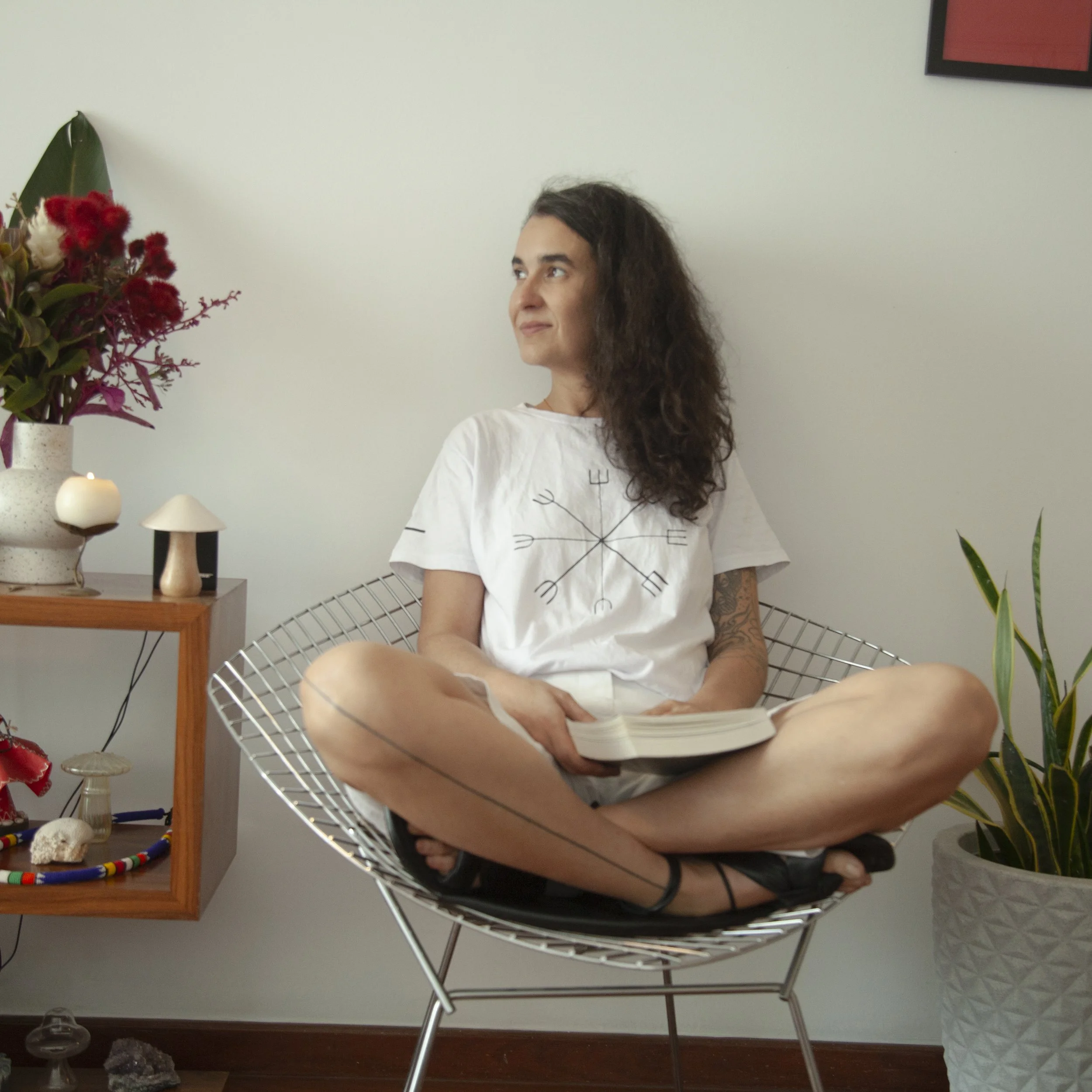 A woman with curly hair sitting cross-legged on a wire chair, reading a book in a room decorated with plants, candles, and artwork.