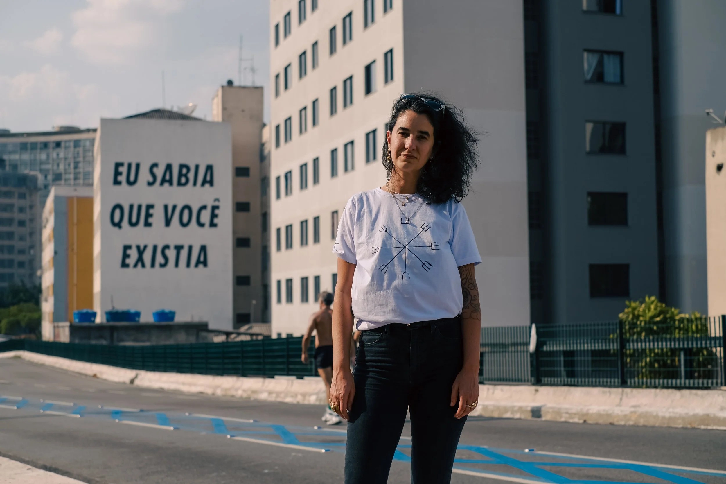 A woman with dark hair and tattoos on her arm standing in an urban area. Behind her is a large building with a sign that reads, 'EU SABIA QUE VOCE EXISTIA'.