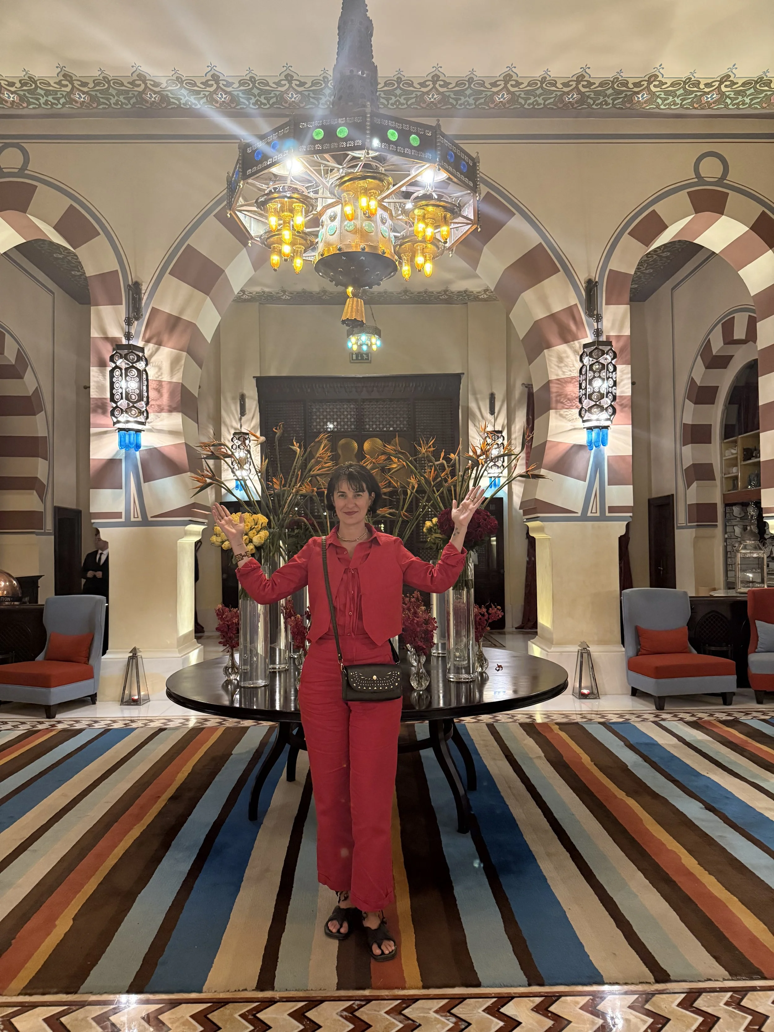 A woman in a red outfit stands in a luxurious hotel lobby with colorful lights, large floral arrangements, and a striped carpet.